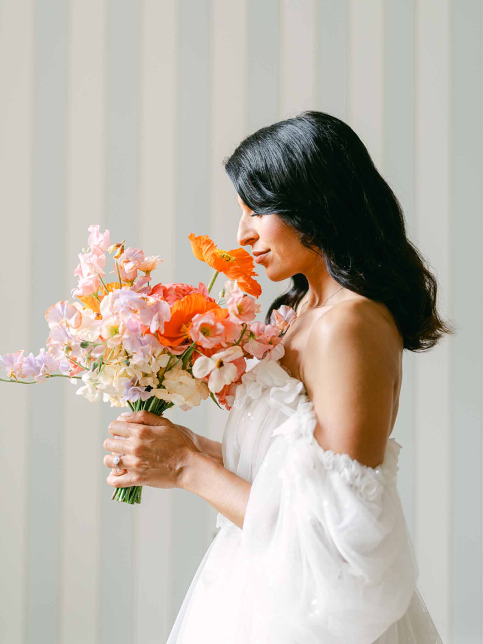 Bride in off-the-shoulder white dress smelling a garden-style bouquet of orange poppies and sweet peas