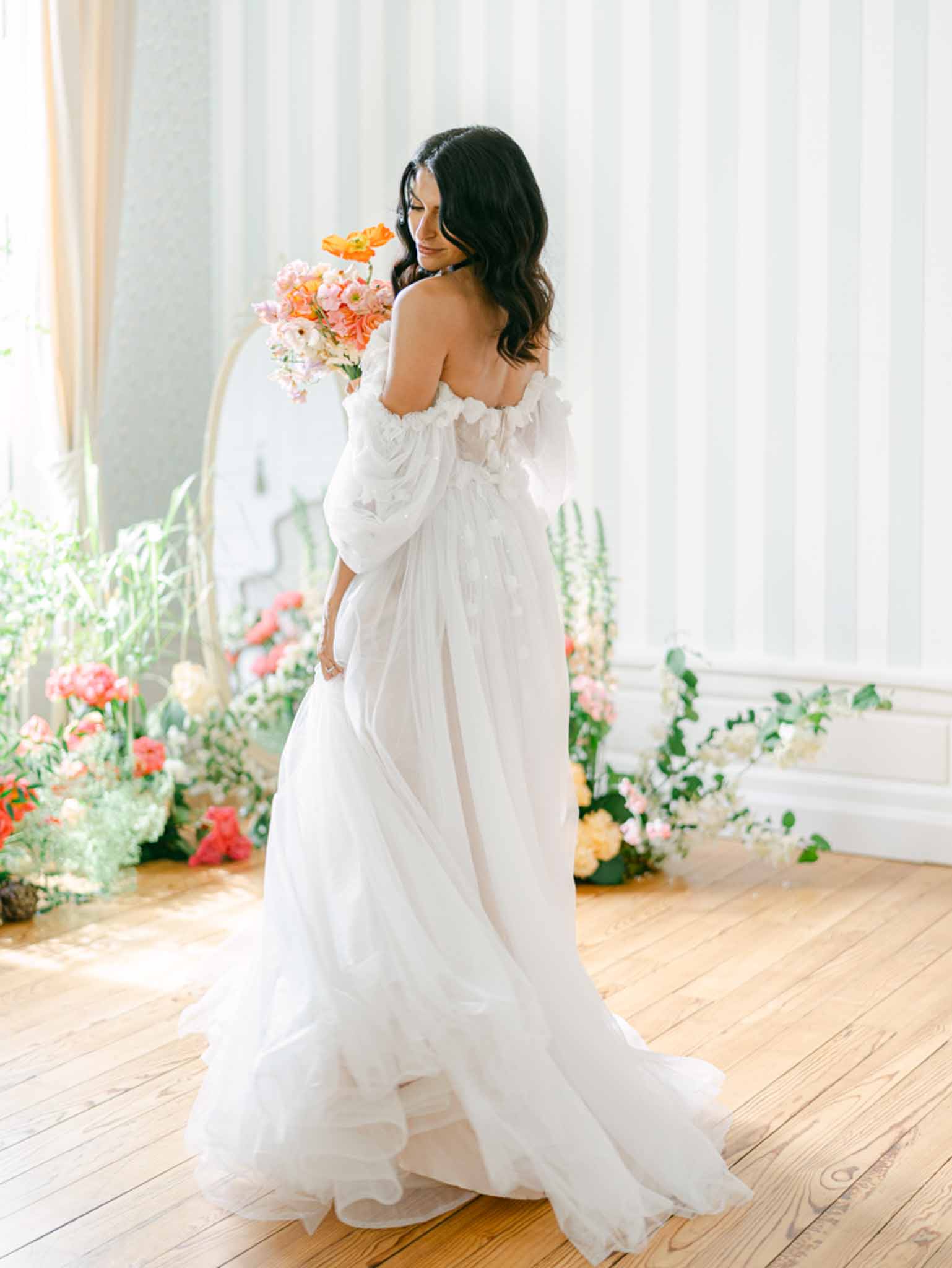 Bride in off-the-shoulder white tulle ball gown holding garden bouquet of orange poppies and coral ranunculus indoors