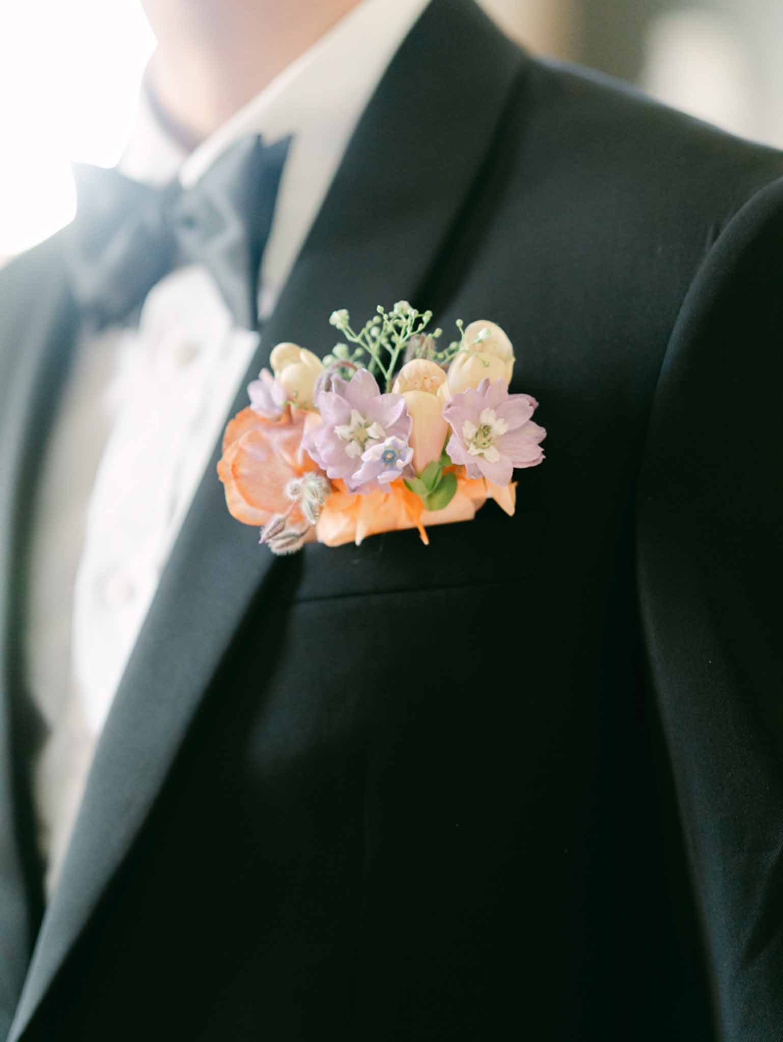Groom boutonniere with peach rose and lavender delphinium pinned to black tuxedo lapel with bow tie