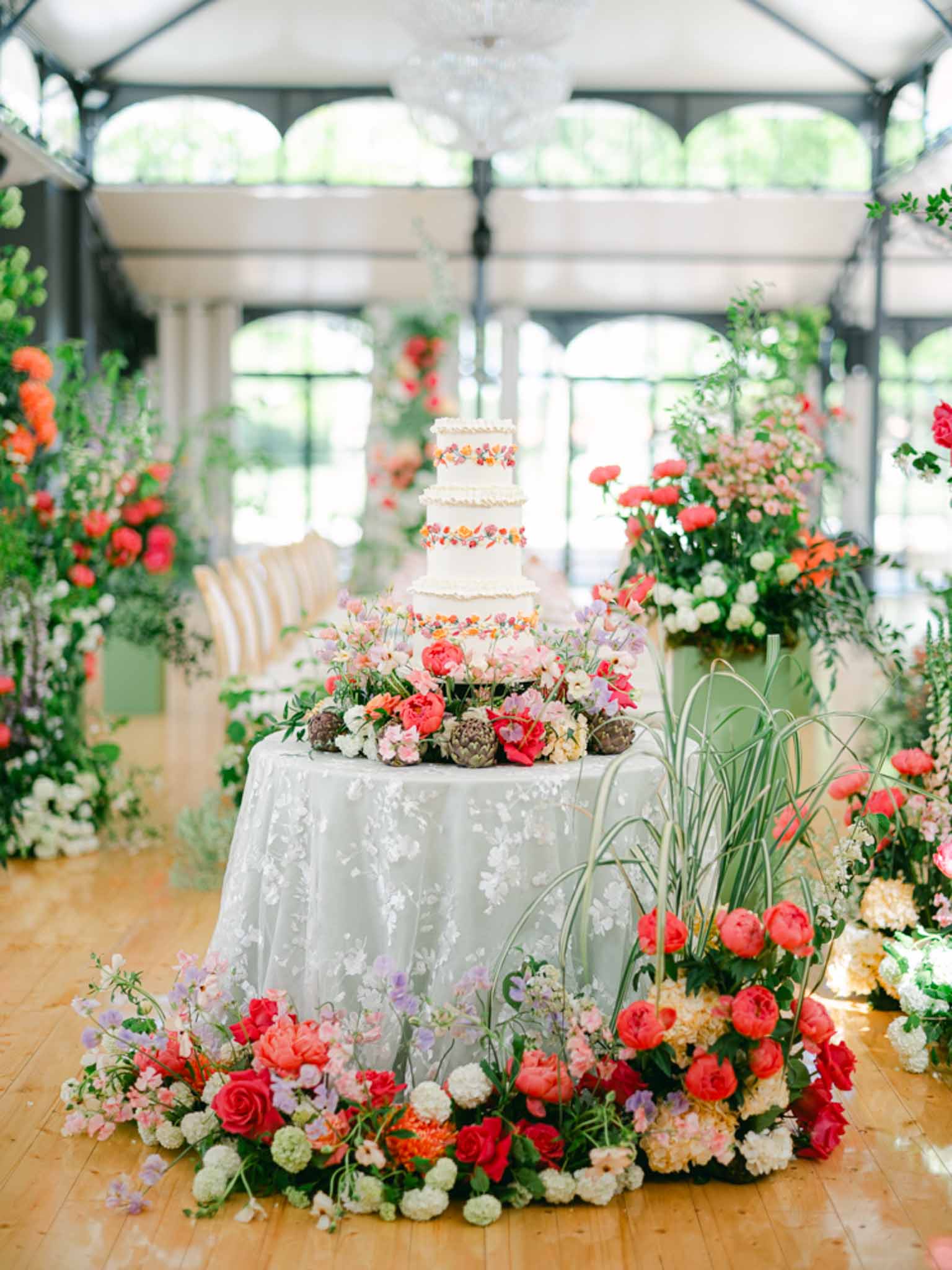 Four-tier painted floral cake surrounded by coral peony and rose columns in glass-and-iron greenhouse with chandelier
