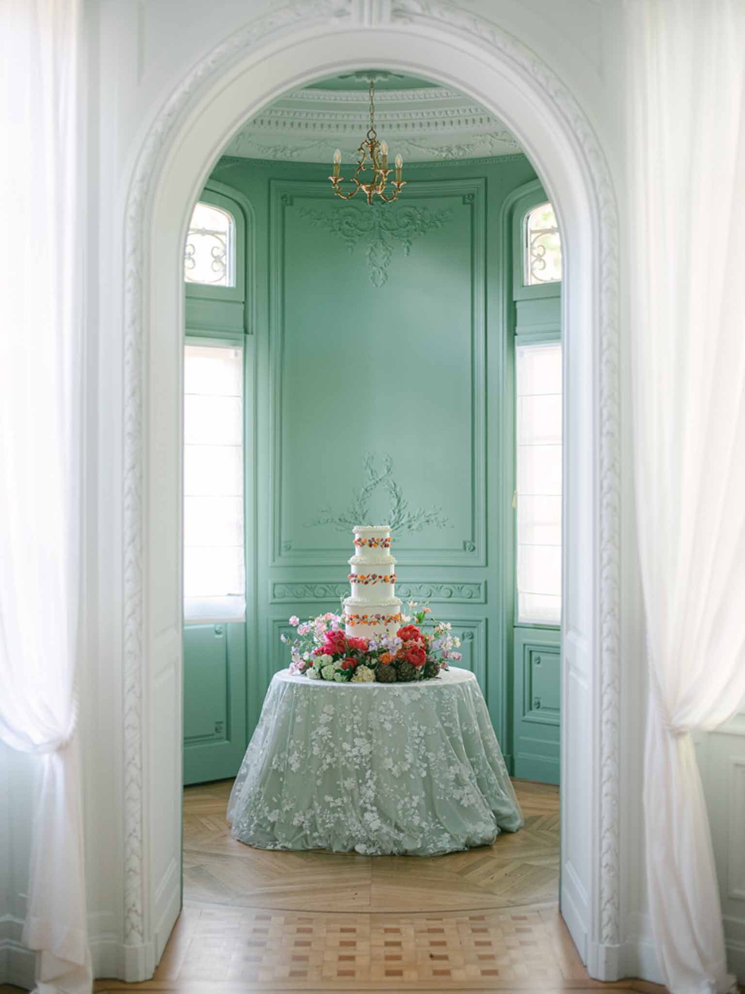 Four-tier white cake surrounded by colorful blooms in sage green paneled alcove with brass chandelier