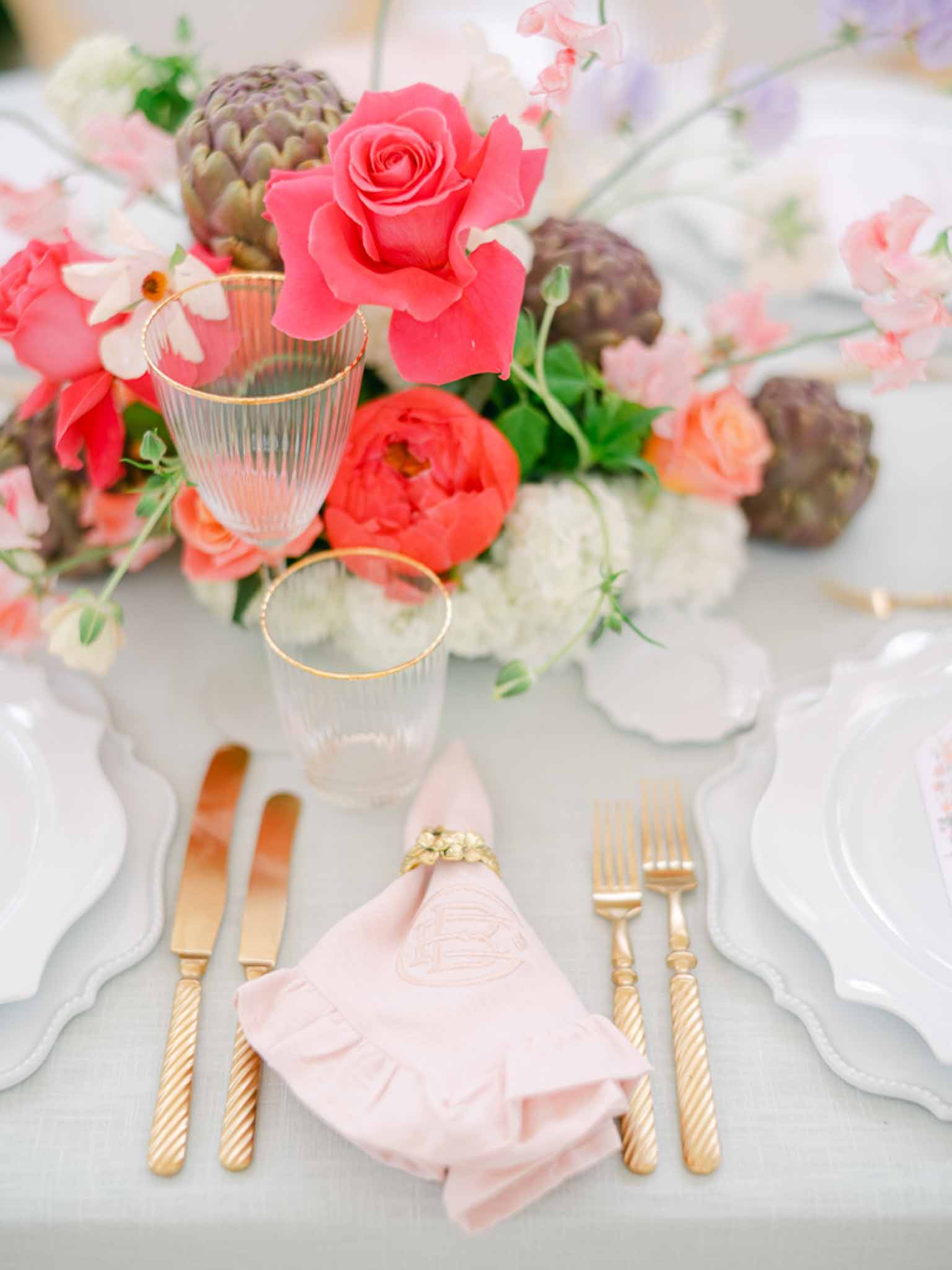 Place setting with scallop charger, gold flatware, blush embroidered napkin, and coral rose centerpiece