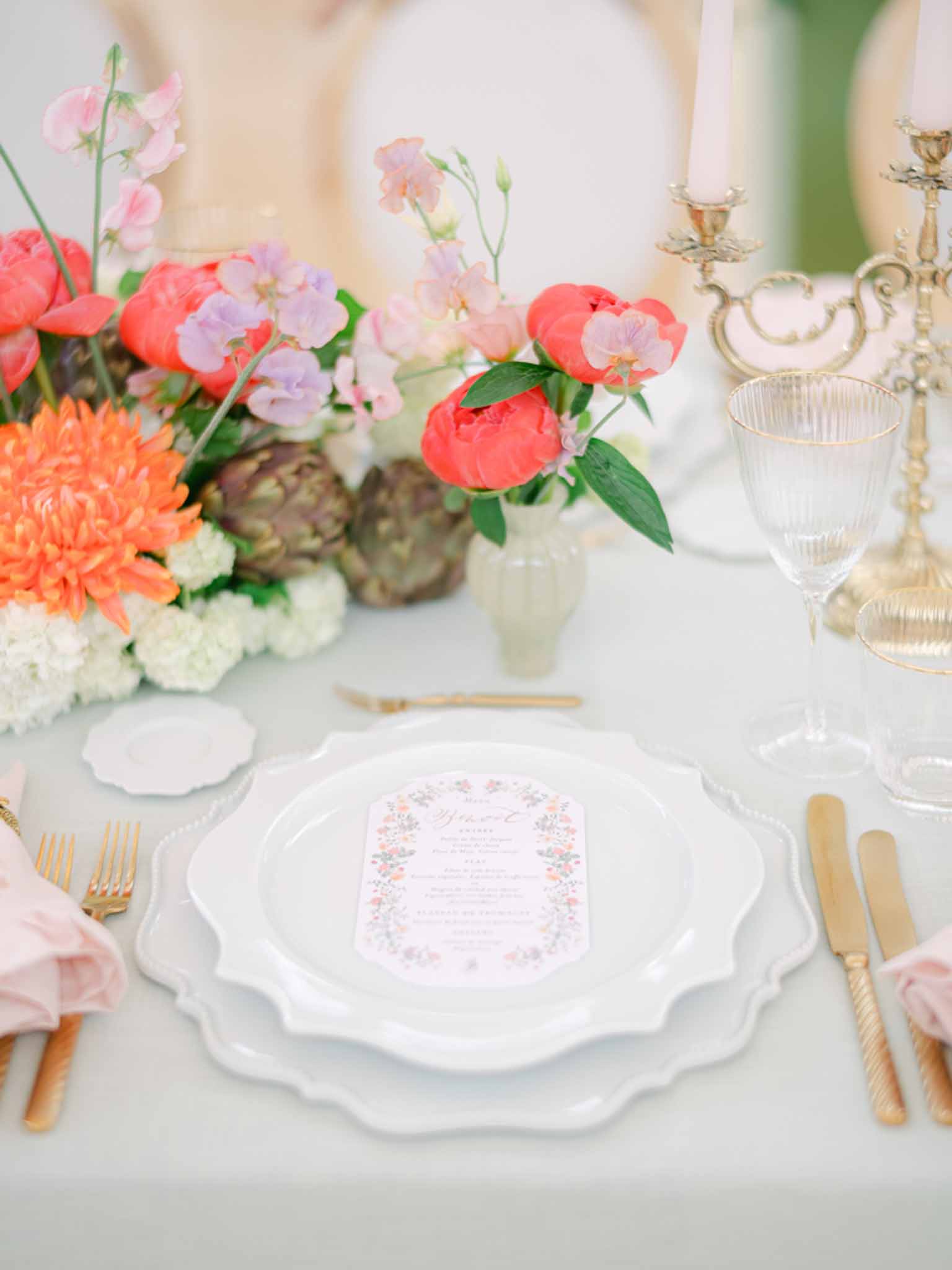 Wedding reception place setting with scallop-edged plates, printed menu card, gold flatware, and coral peony centerpiece