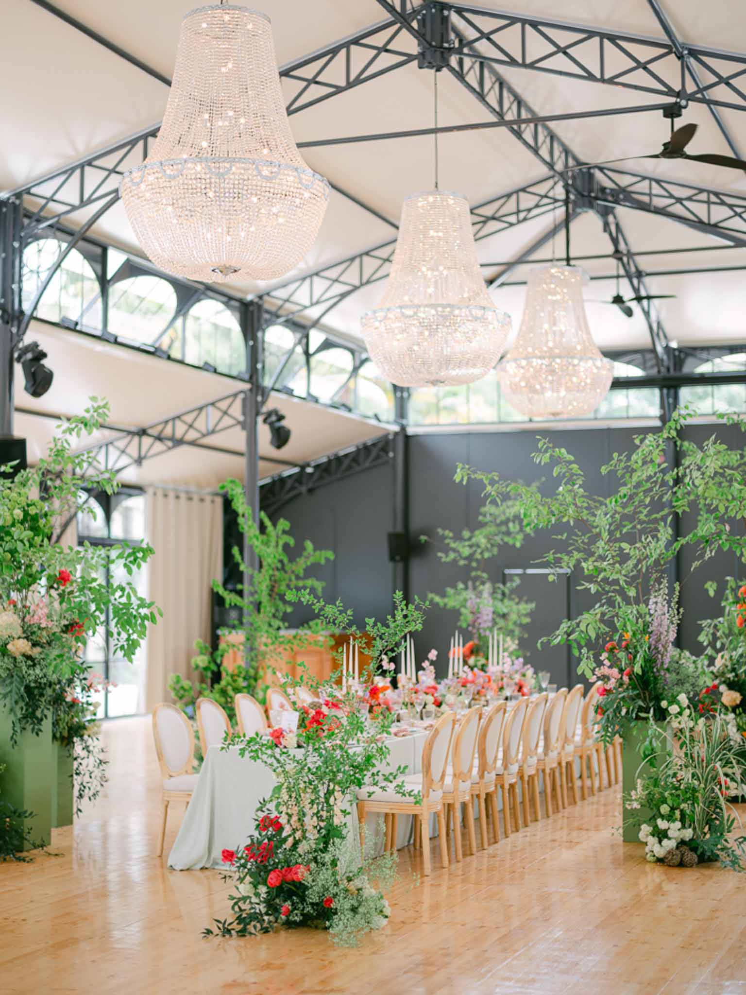 Long feasting table under crystal chandeliers in a greenhouse venue with coral and red floral arrangements