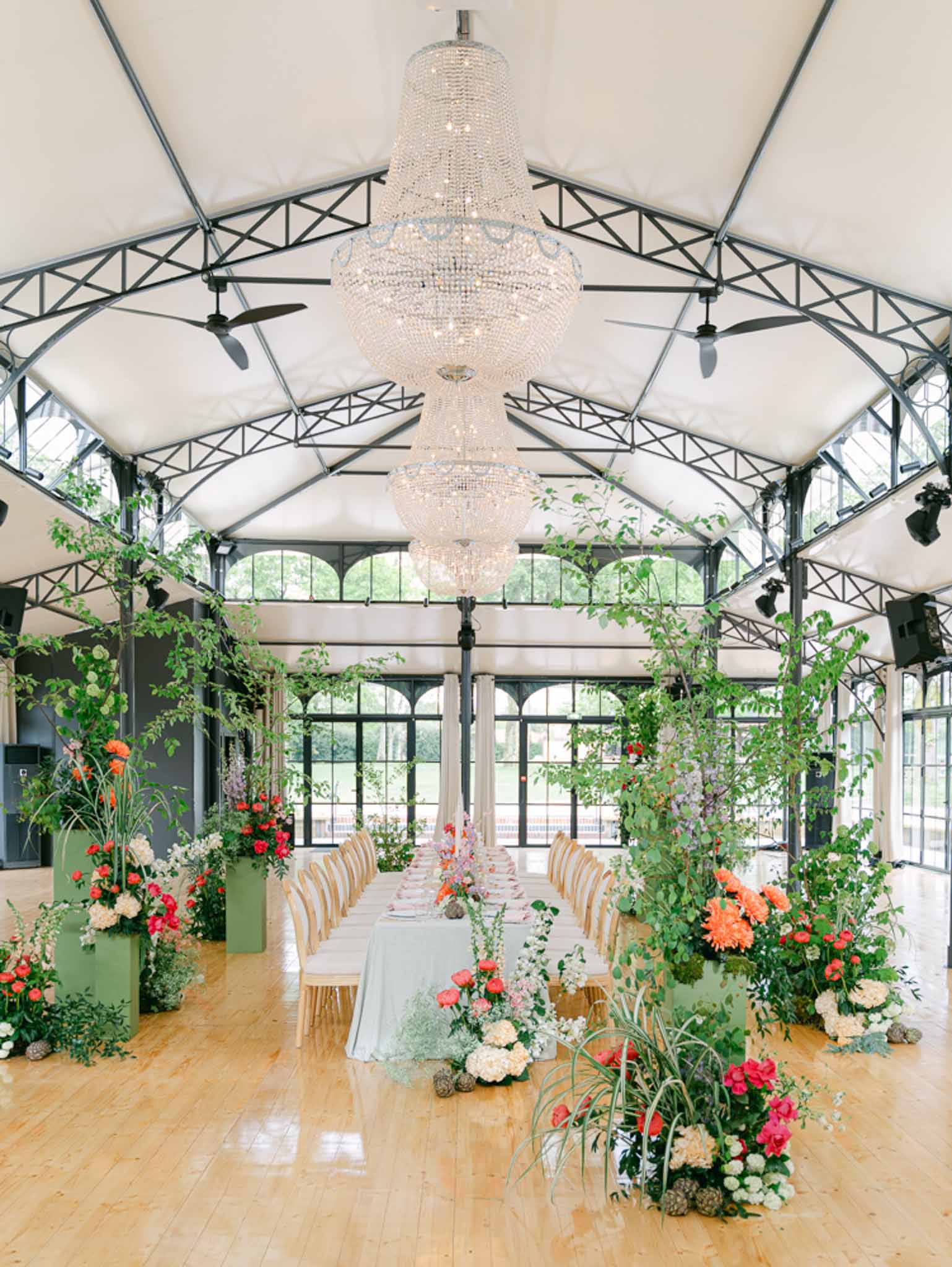 Greenhouse wedding reception with long banquet table, sage green linens, crystal chandeliers, and lush coral floral displays