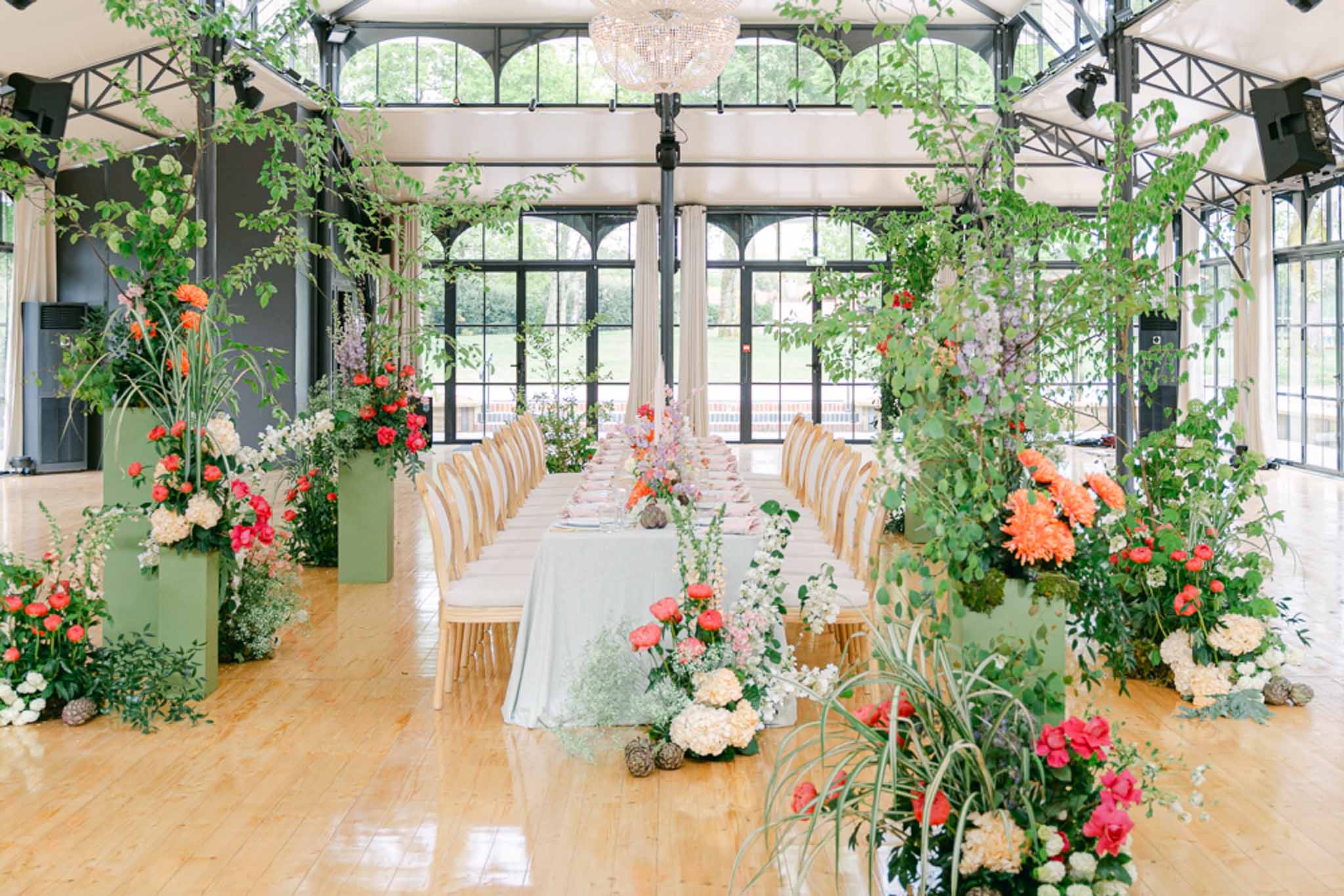 Pavilion reception with sage plinths, coral ranunculus, crystal chandelier, and climbing branches overhead