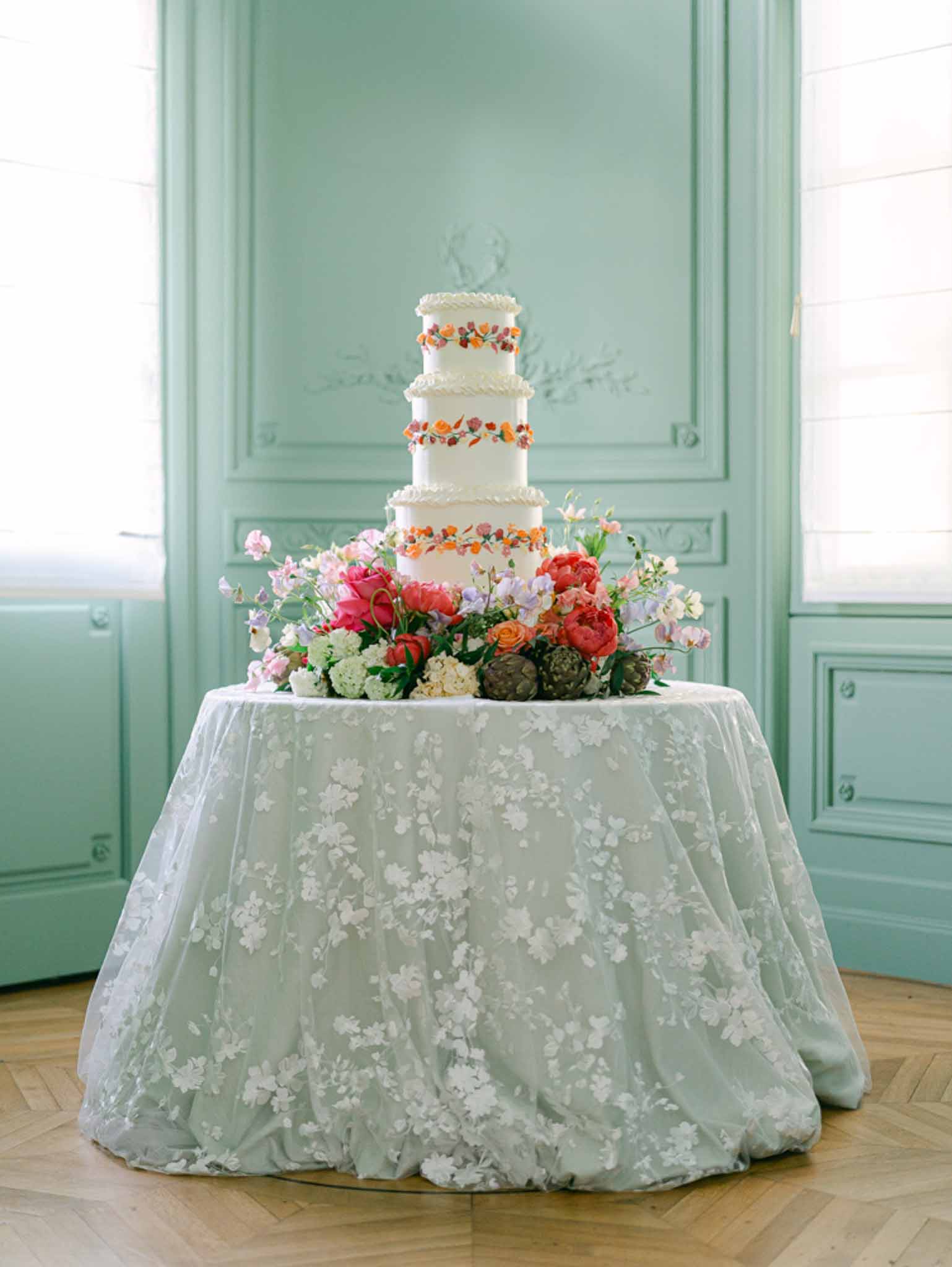 Four-tier white ruffle cake with coral sugar flowers beside hot pink peonies on blue-grey jacquard linen
