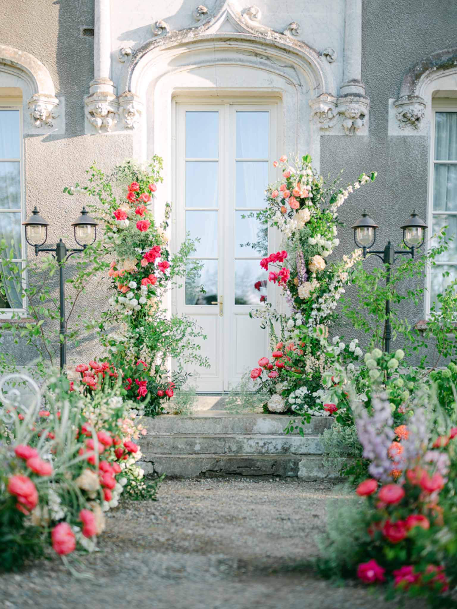 Chateau entrance with tall floral columns of coral roses, white hydrangeas, and lavender delphiniums framing white double ...