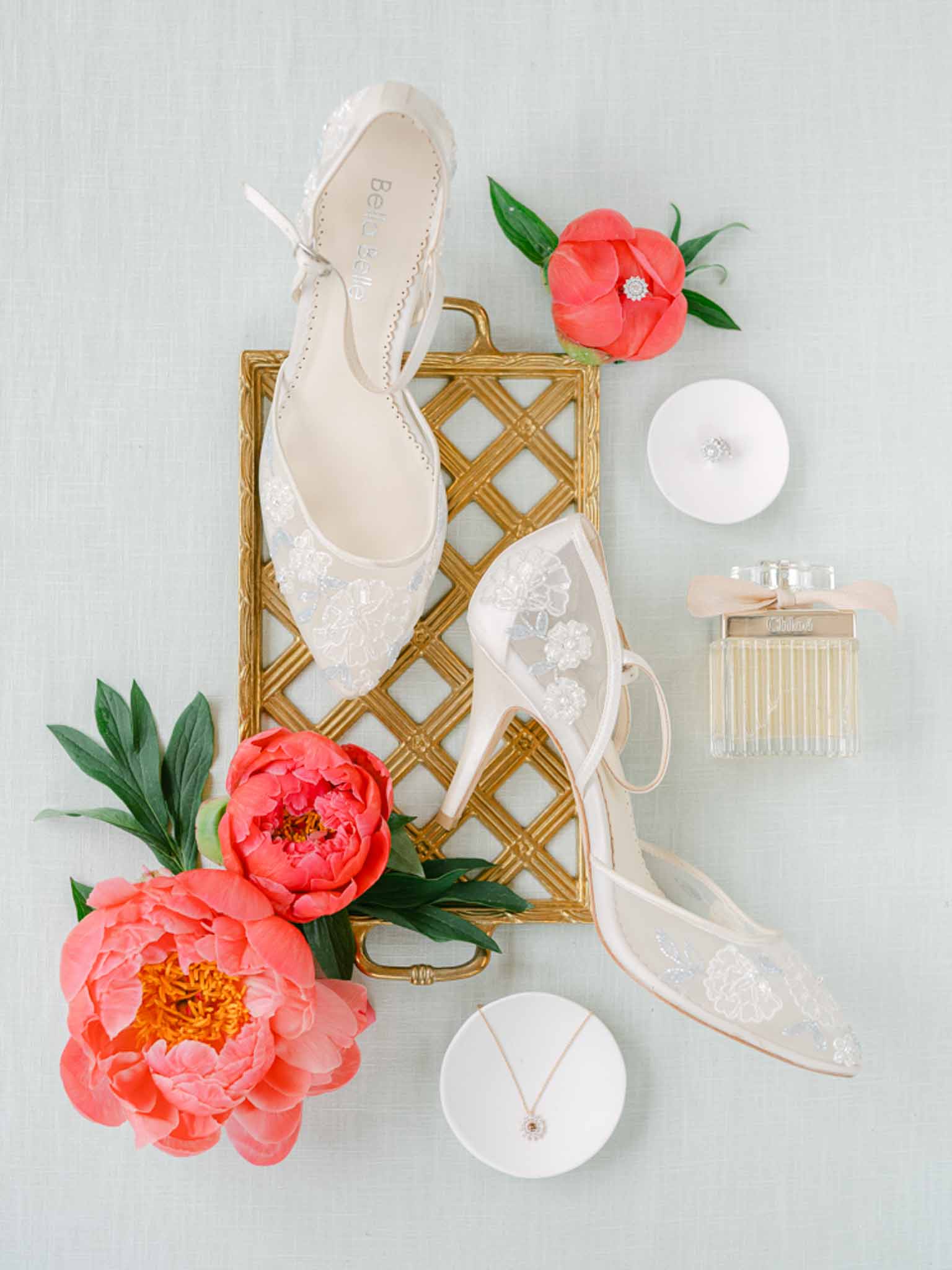 Bridal flat lay with ivory lace Bella Belle heels, coral peonies, Chloe perfume, and rose gold jewelry on mint linen