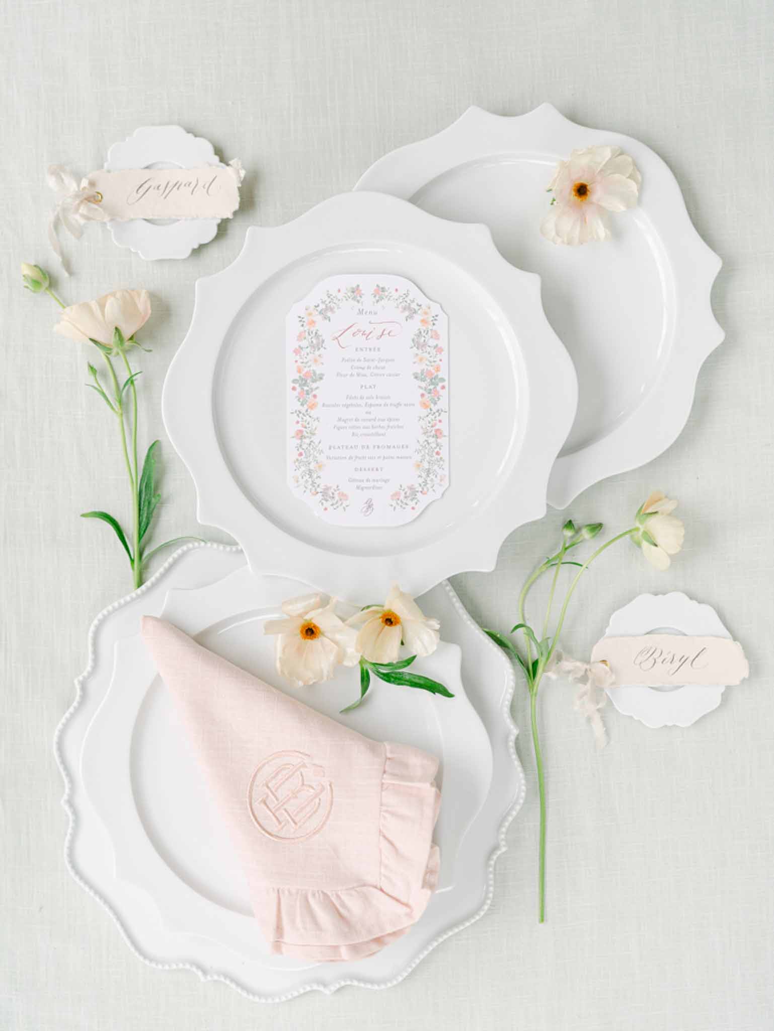 Flat lay of place setting with scallop-edged plates, monogrammed pink napkin, menu card, and ranunculus blooms