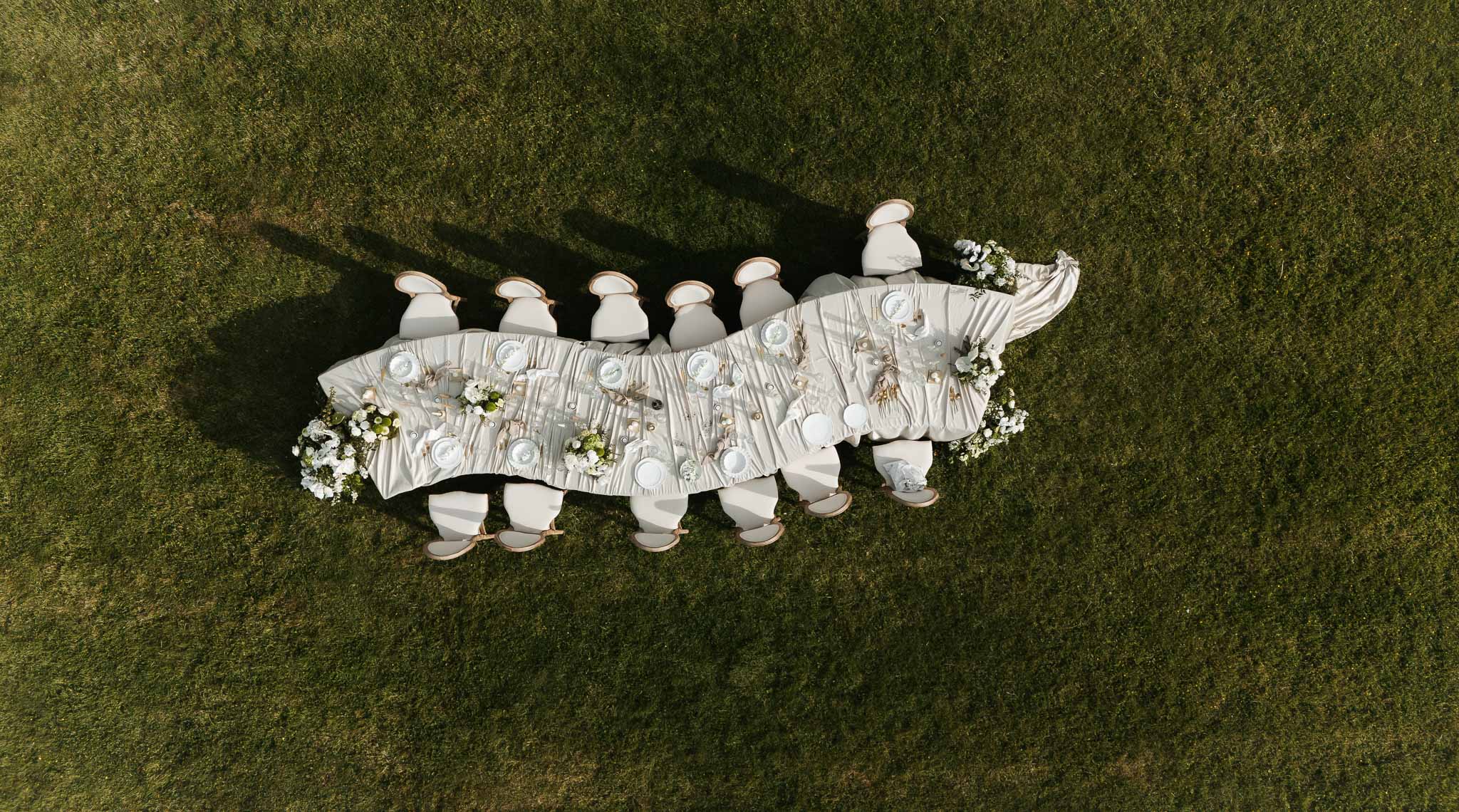 Aerial view of curved outdoor reception table with draped ivory runner, upholstered chairs, and white hydrangeas