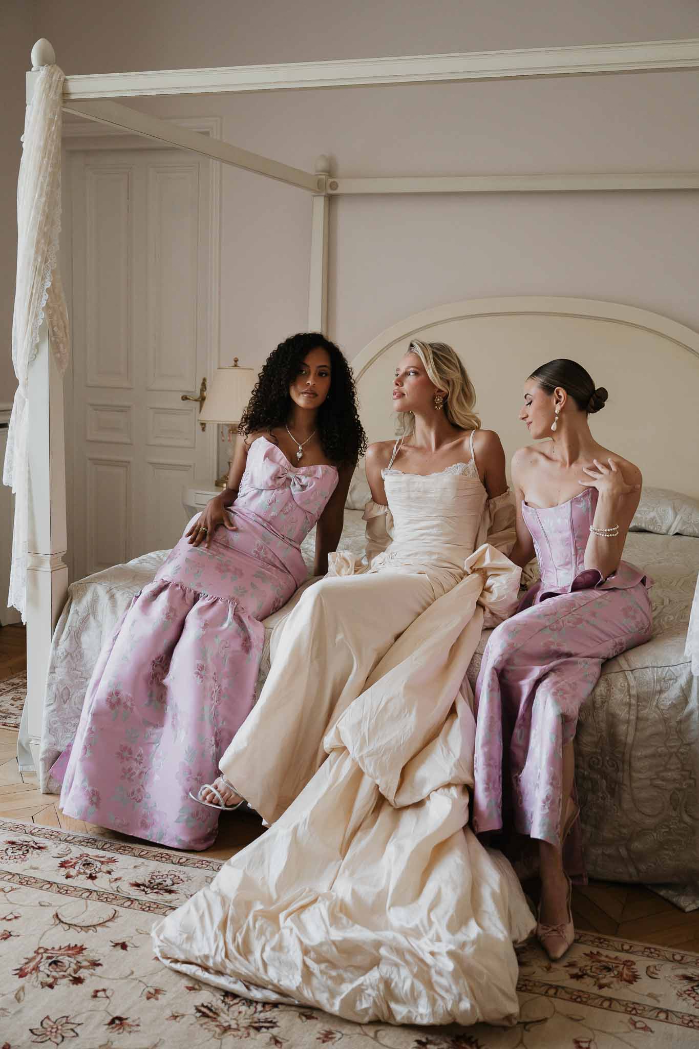 Bride in ivory silk ballgown seated on white four-poster bed with two bridesmaids in mauve floral gowns