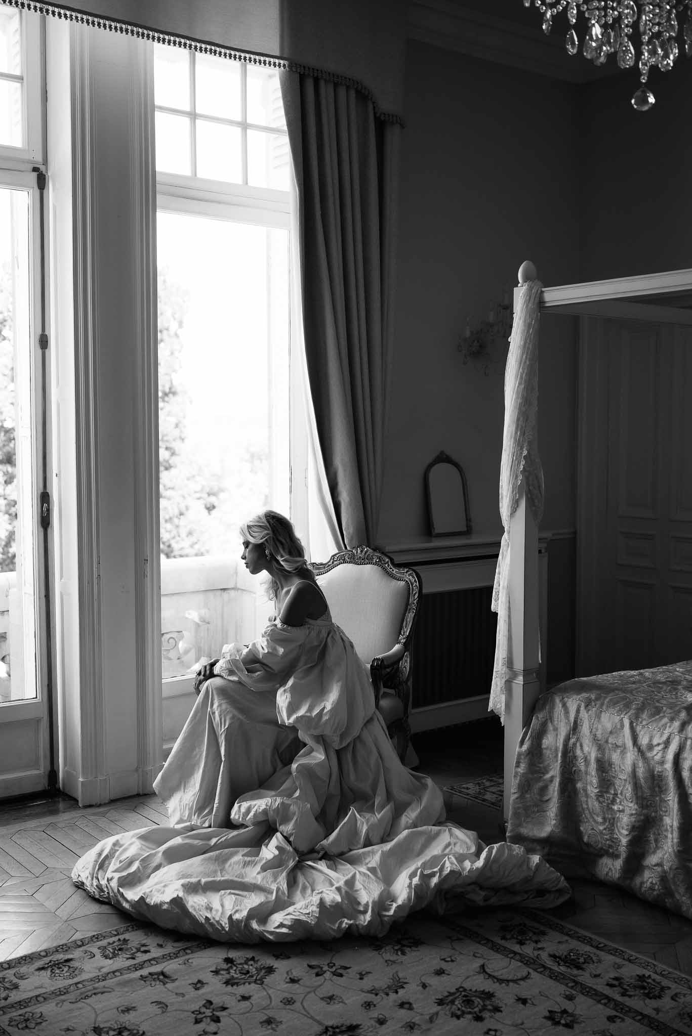 Black-and-white portrait of bride in off-shoulder gown seated by French windows in ornate chateau room