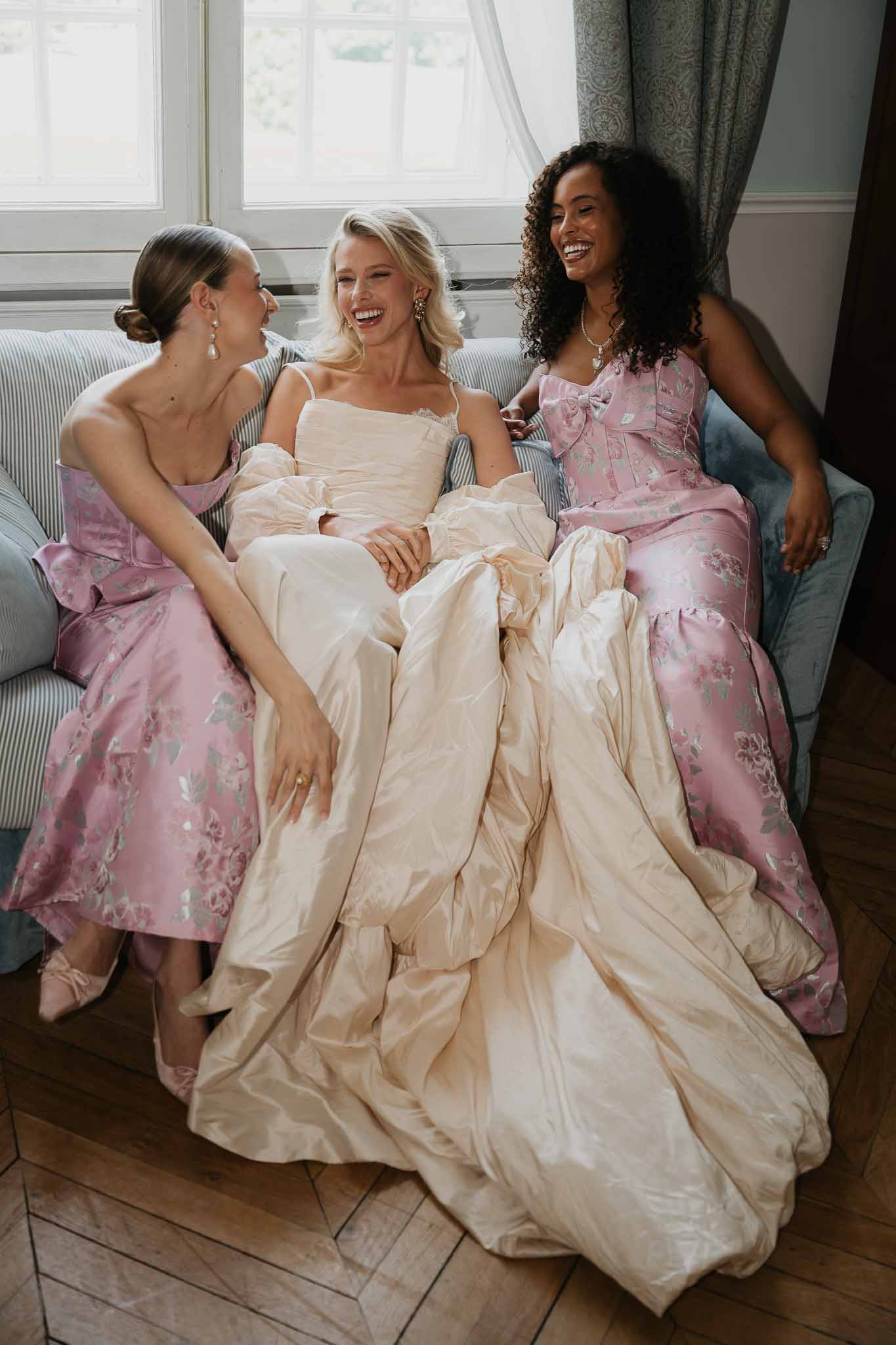 Bride in champagne ball gown laughing between two bridesmaids in mauve floral jacquard on ticking-stripe sofa