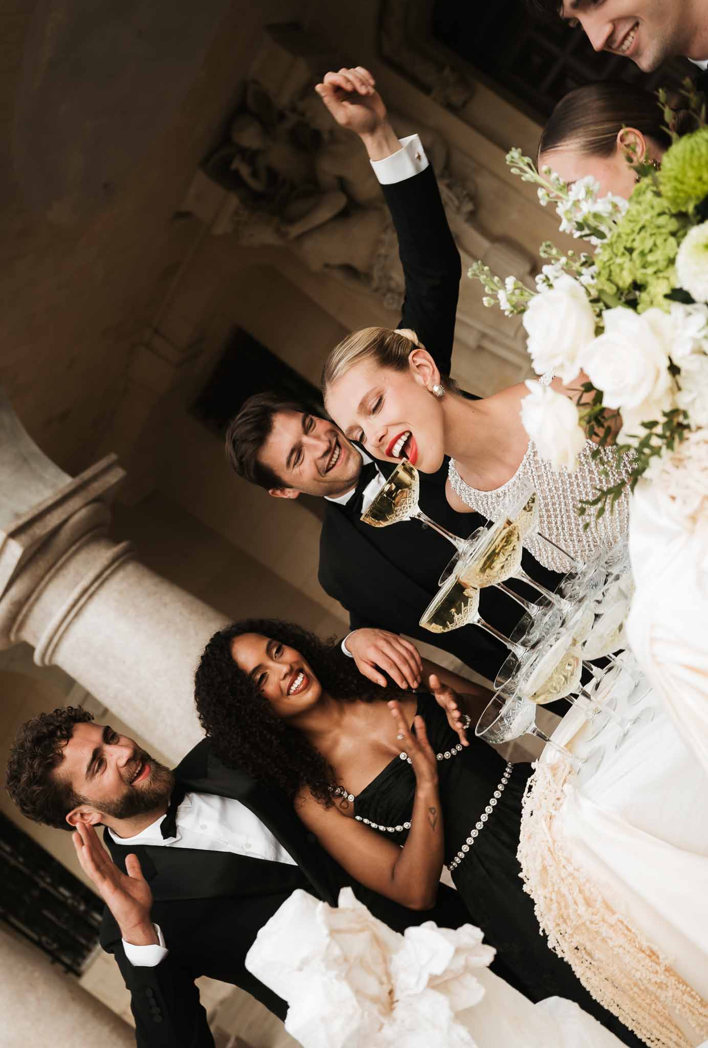 Bride and groom with guests laughing around a champagne coupe tower inside an ornate French chateau salon