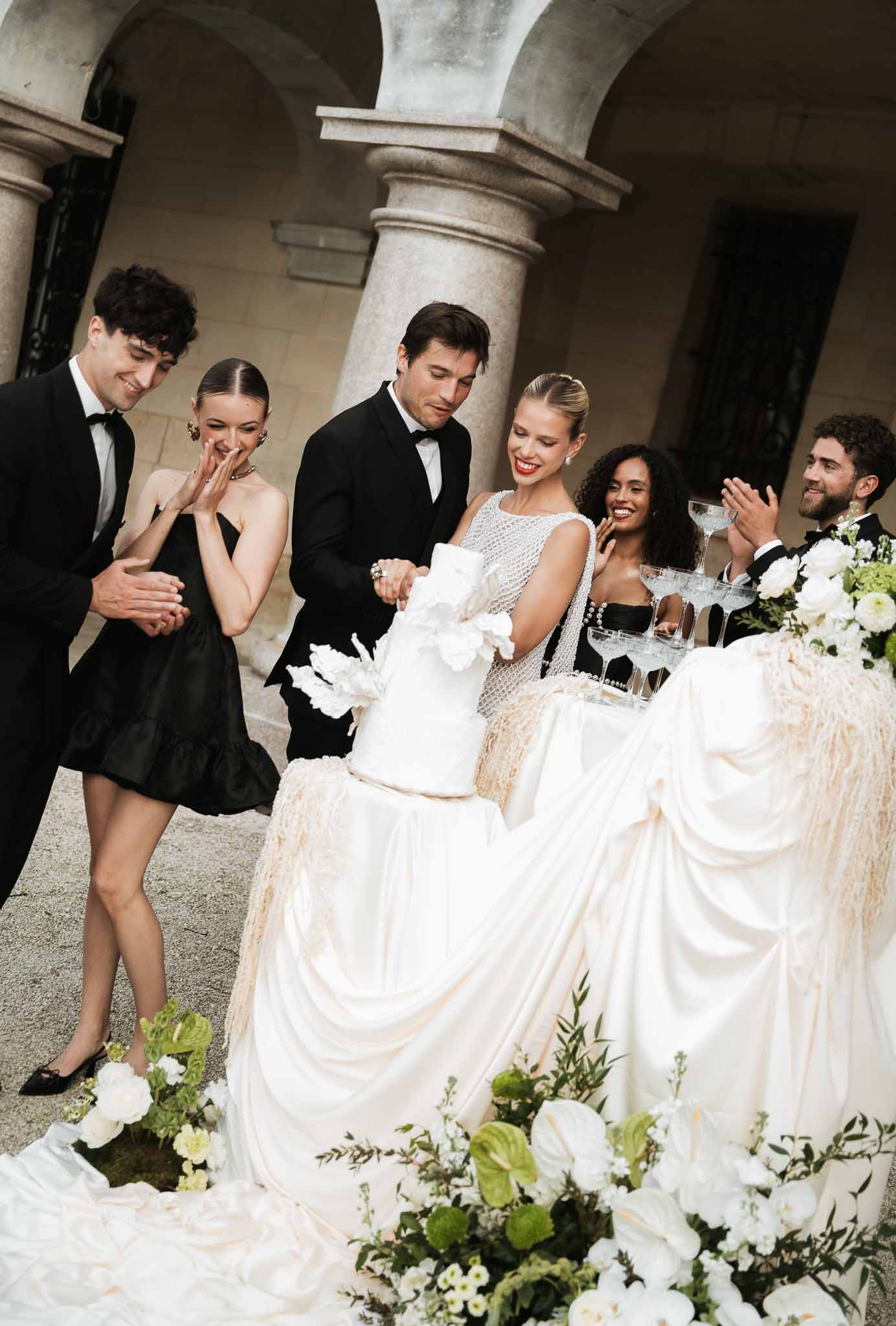 Bride and groom cutting three-tier white cake in stone colonnade courtyard with guests applauding