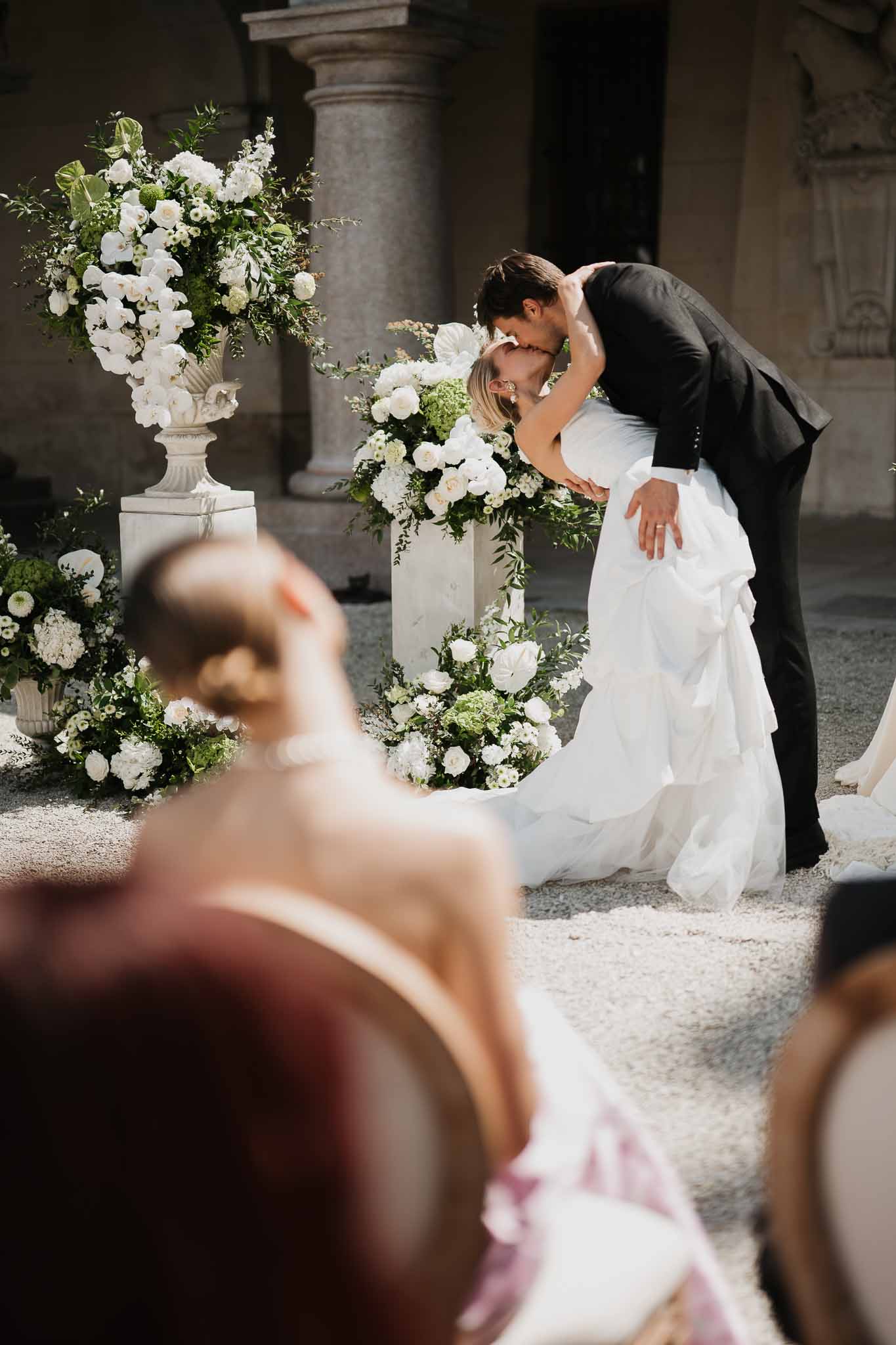 Dip kiss at stone column altar with white orchid and hydrangea urns and ground-level floral clusters