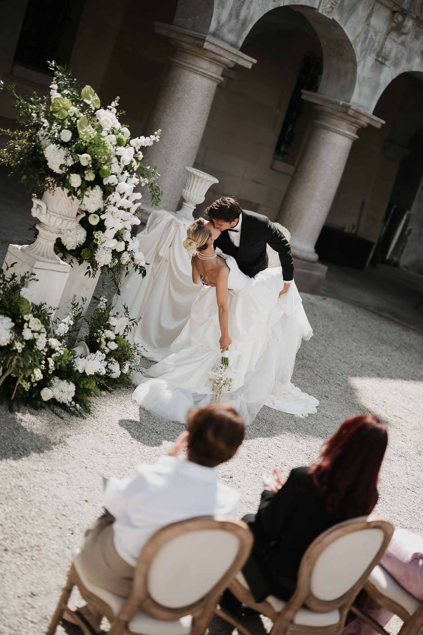 First kiss at stone colonnade altar with white urn pedestals and rose, hydrangea, and delphinium arrangements