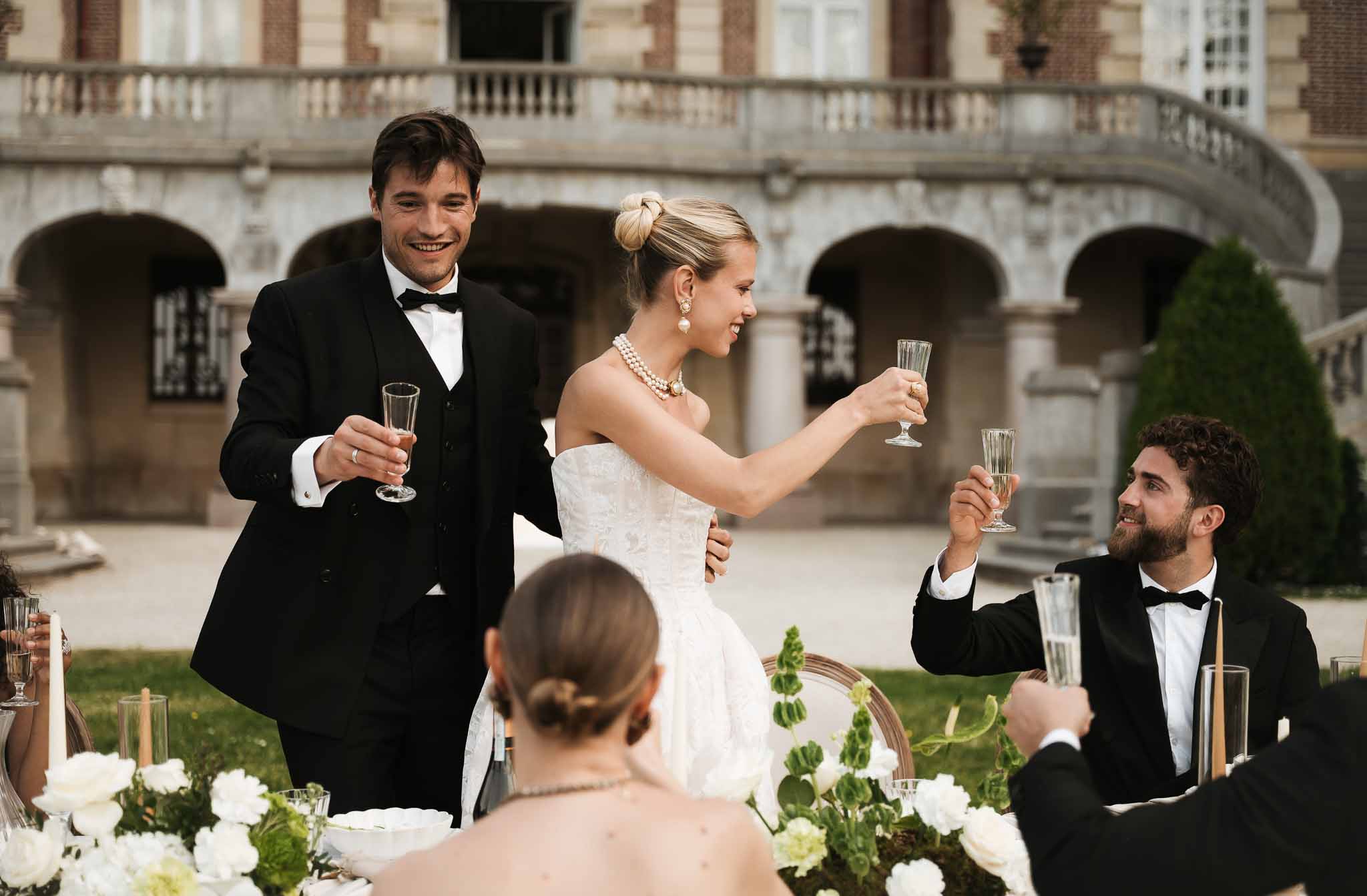 Couple raising champagne flutes with guests at candlelit table before chateau stone colonnades