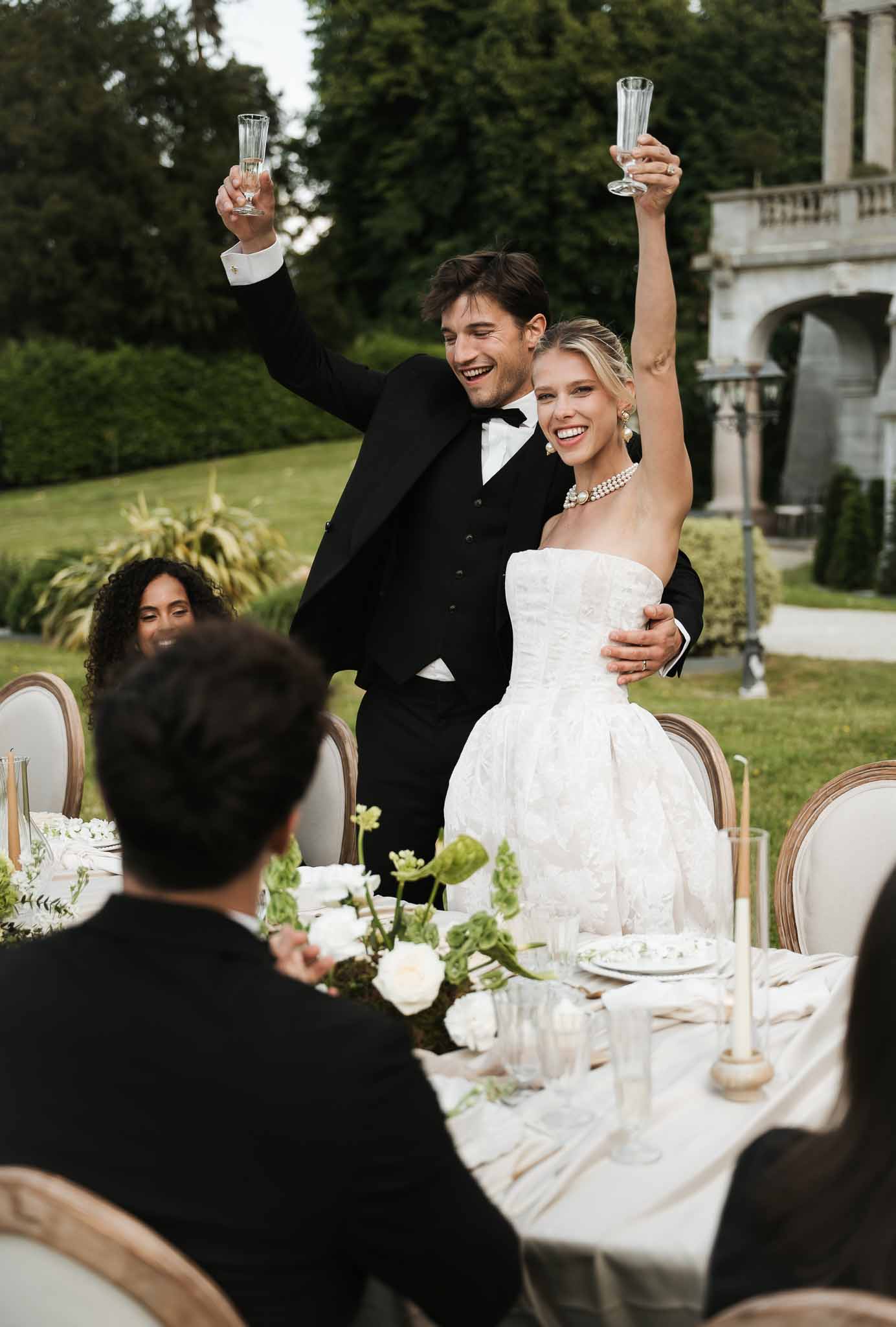Couple raising champagne flutes at outdoor reception with white rose centerpieces and stone balustrade behind