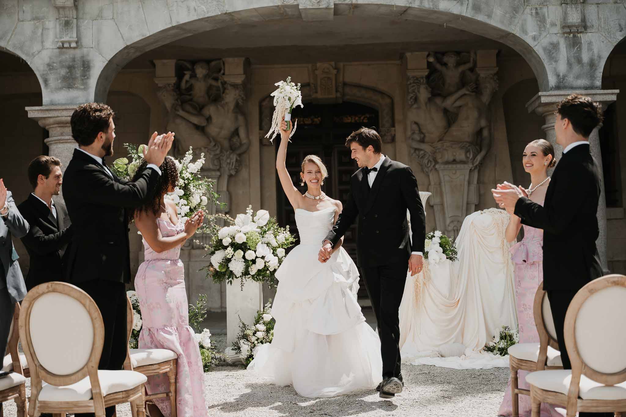 Couple recessional through colonnade courtyard with bride raising bouquet, flanked by groomsmen and pink-gown bridesmaids