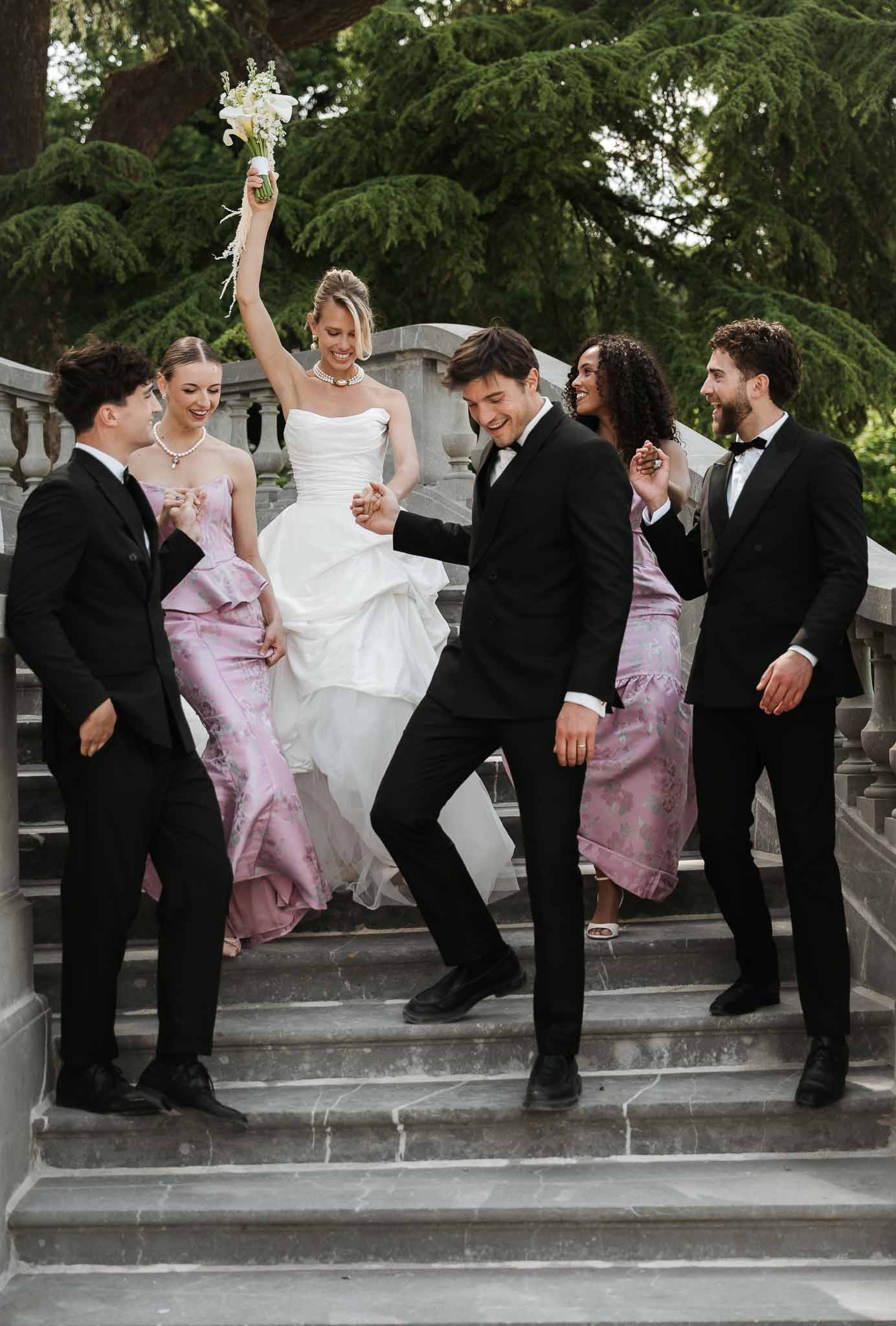 Bridal party of six laughing on stone steps, bridesmaids in mauve-pink jacquard gowns, groomsmen in black tuxedos