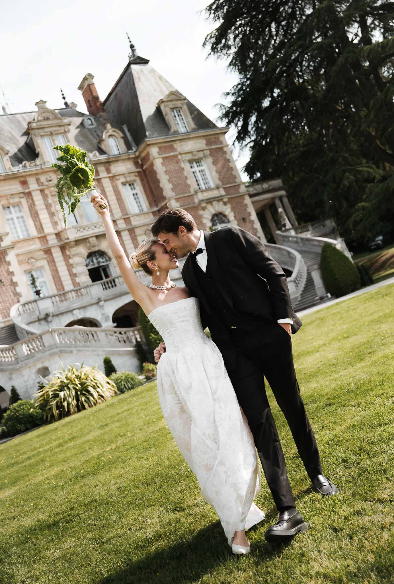 Bride in strapless gown with green bouquet raised and groom in black tuxedo nearly kissing on chateau lawn