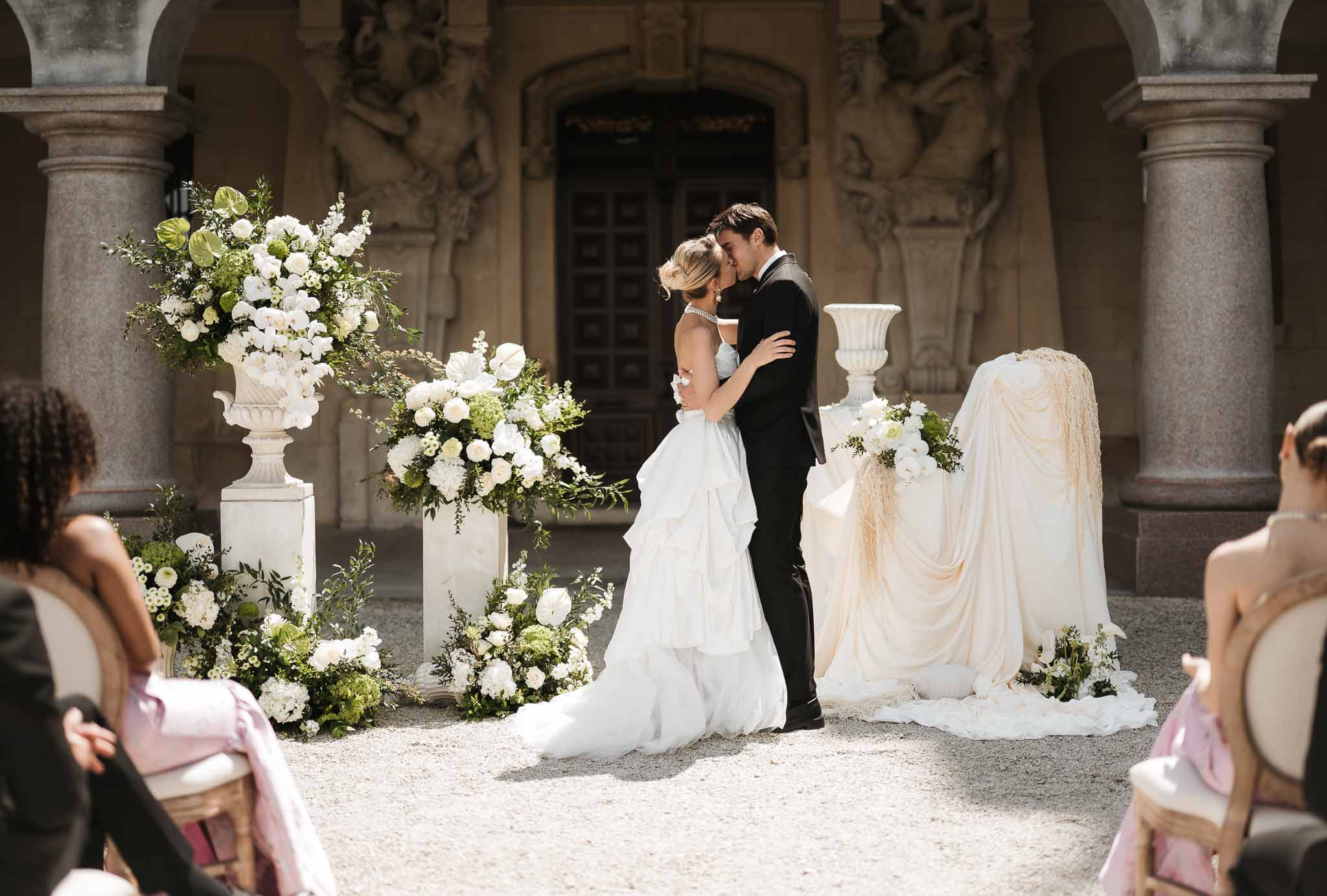 Bride and groom share first kiss at outdoor courtyard ceremony with white rose urns and gold guest chairs