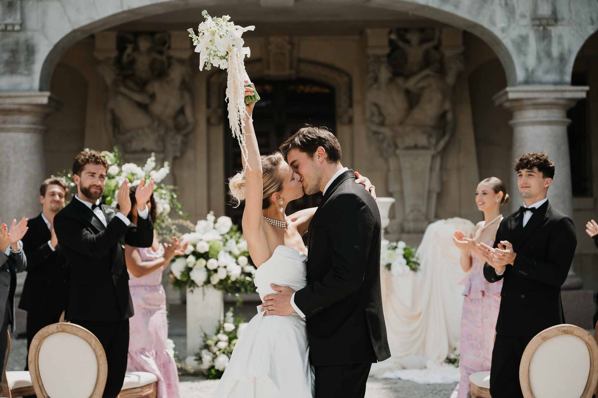 First kiss at ornate stone facade with white floral pedestals and bridal party applauding