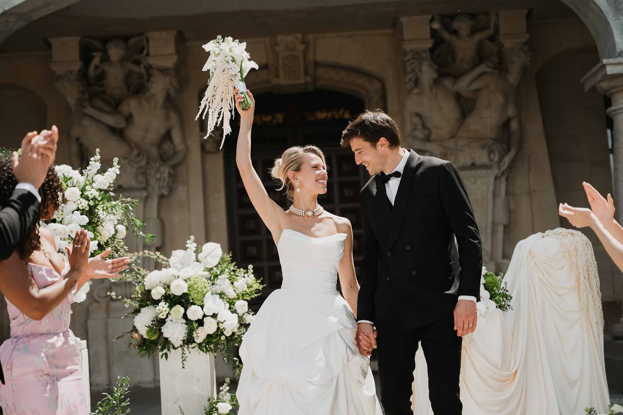 Couple exiting ceremony past white floral pedestals as guests applaud at ornate stone facade