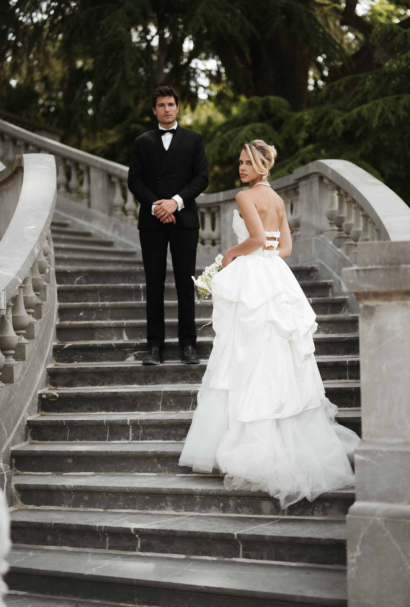 Bride in layered ruffle ballgown with pearl necklace and groom in tuxedo on grand stone staircase