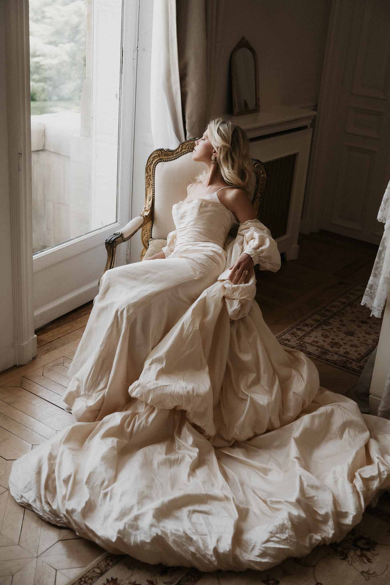 Bride in taffeta ballgown with puff-sleeve jacket reclining in gold Louis XVI chair by chateau window
