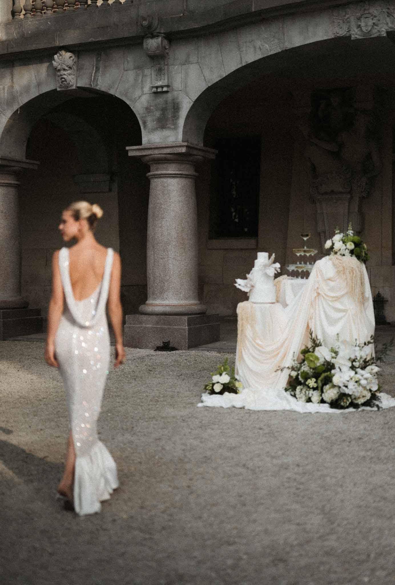 Bride walking toward cake table with champagne tower in chateau courtyard with stone colonnades