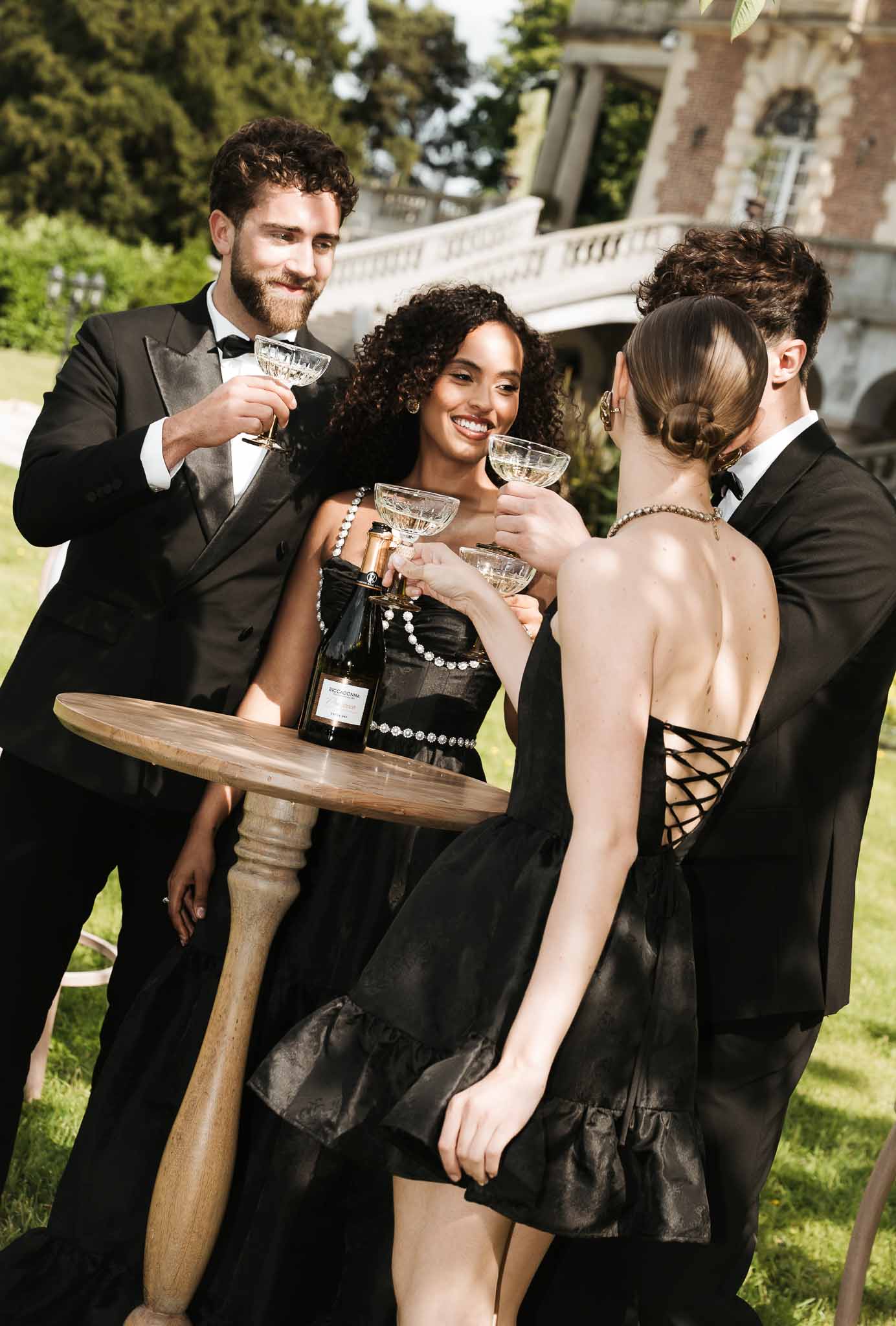 Four guests in black tuxedos and black strapless dresses toasting champagne coupes at round table on chateau terrace