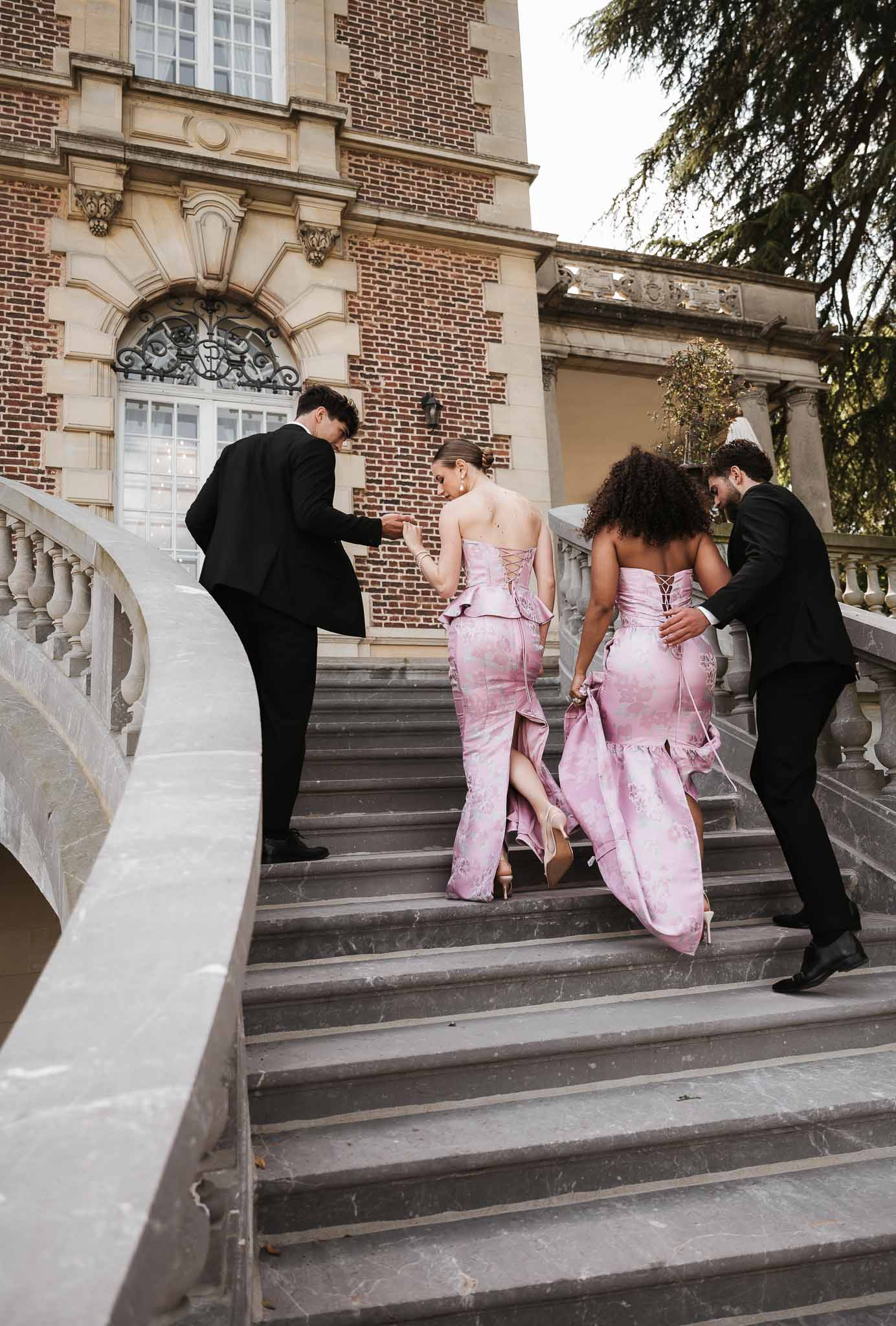 Two bridesmaids in pink floral jacquard gowns escorted up chateau stone staircase by groomsmen