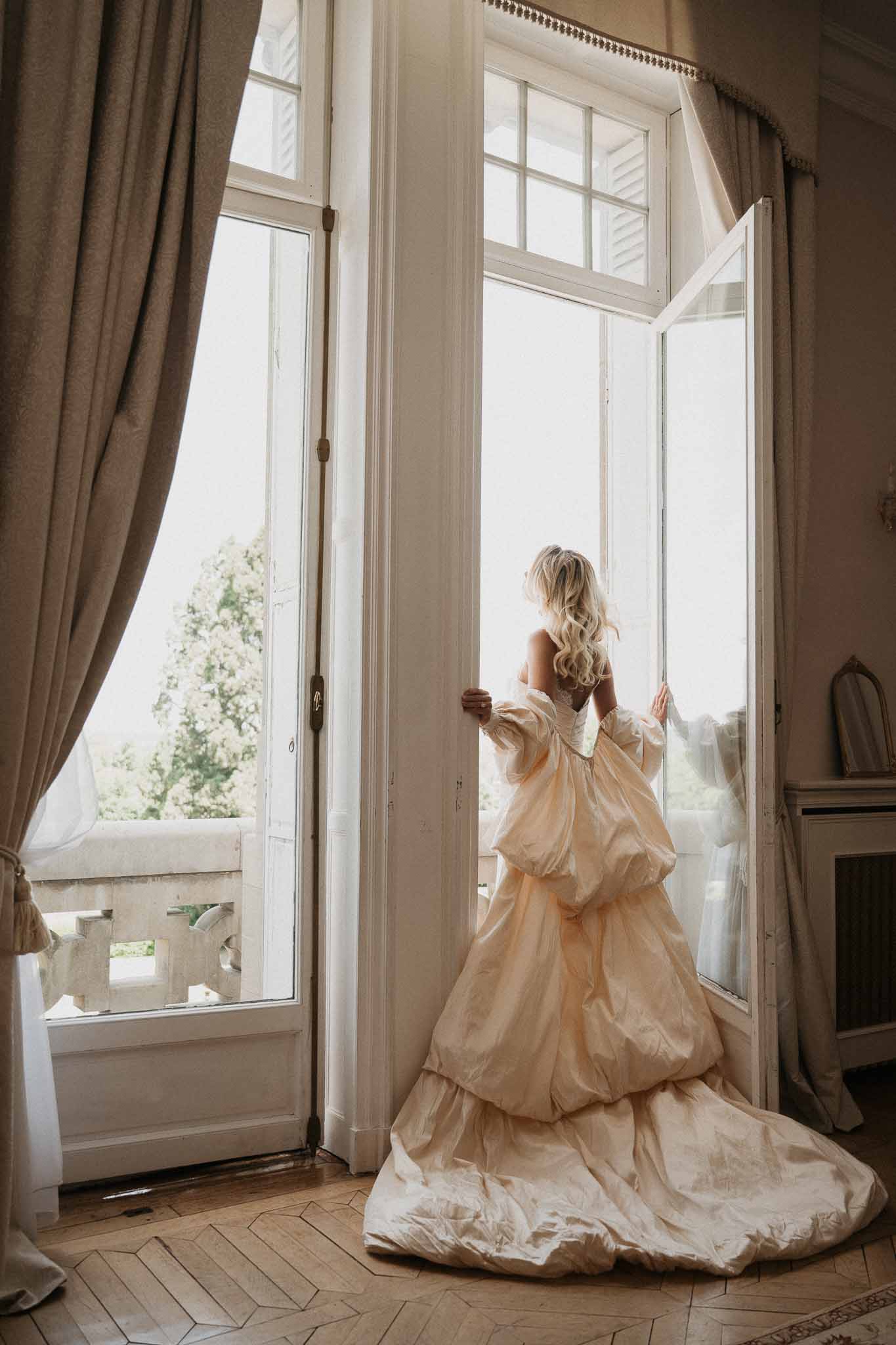 Bride from behind in blush-champagne tiered ballgown with puff sleeves at French doors of chateau getting-ready room