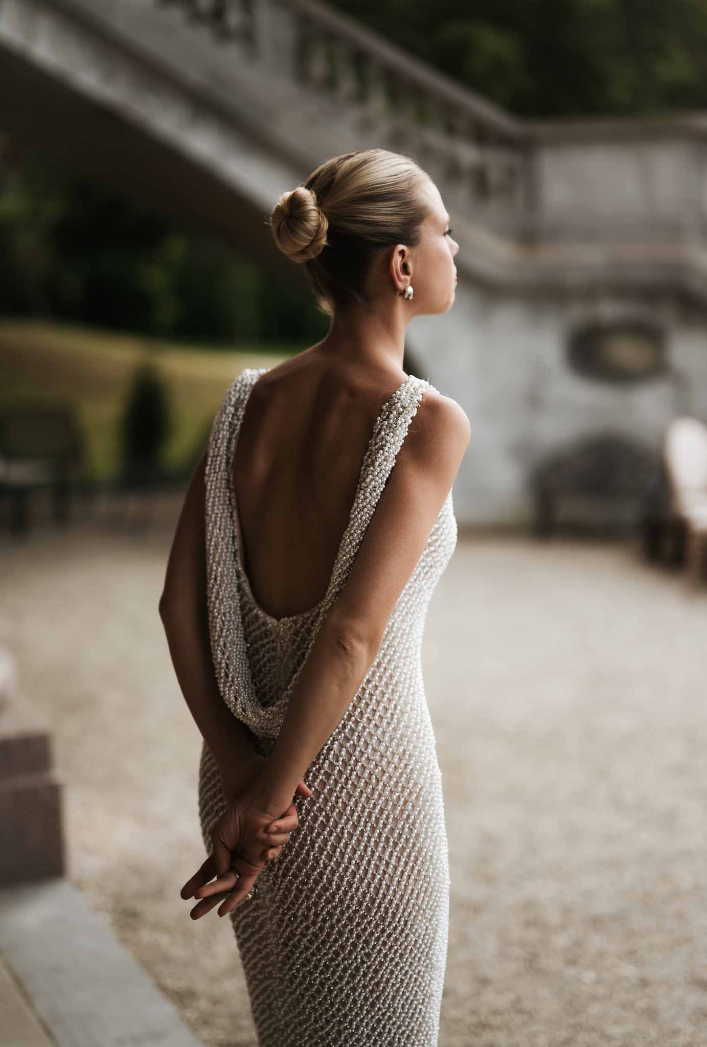 Rear view of bride in pearl-embellished open-back gown with sleek low bun on stone terrace