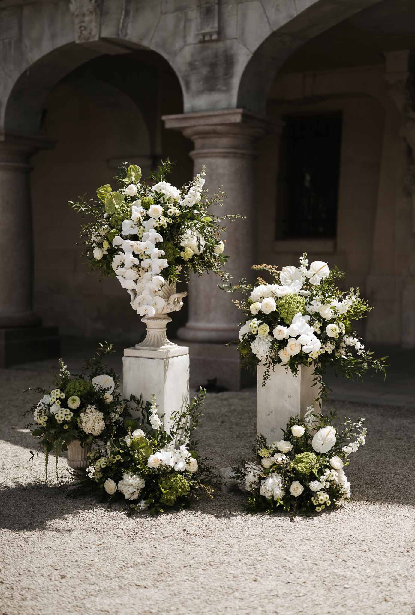 White orchid and rose floral arrangements on marble pedestals in historic venue courtyard with stone arches