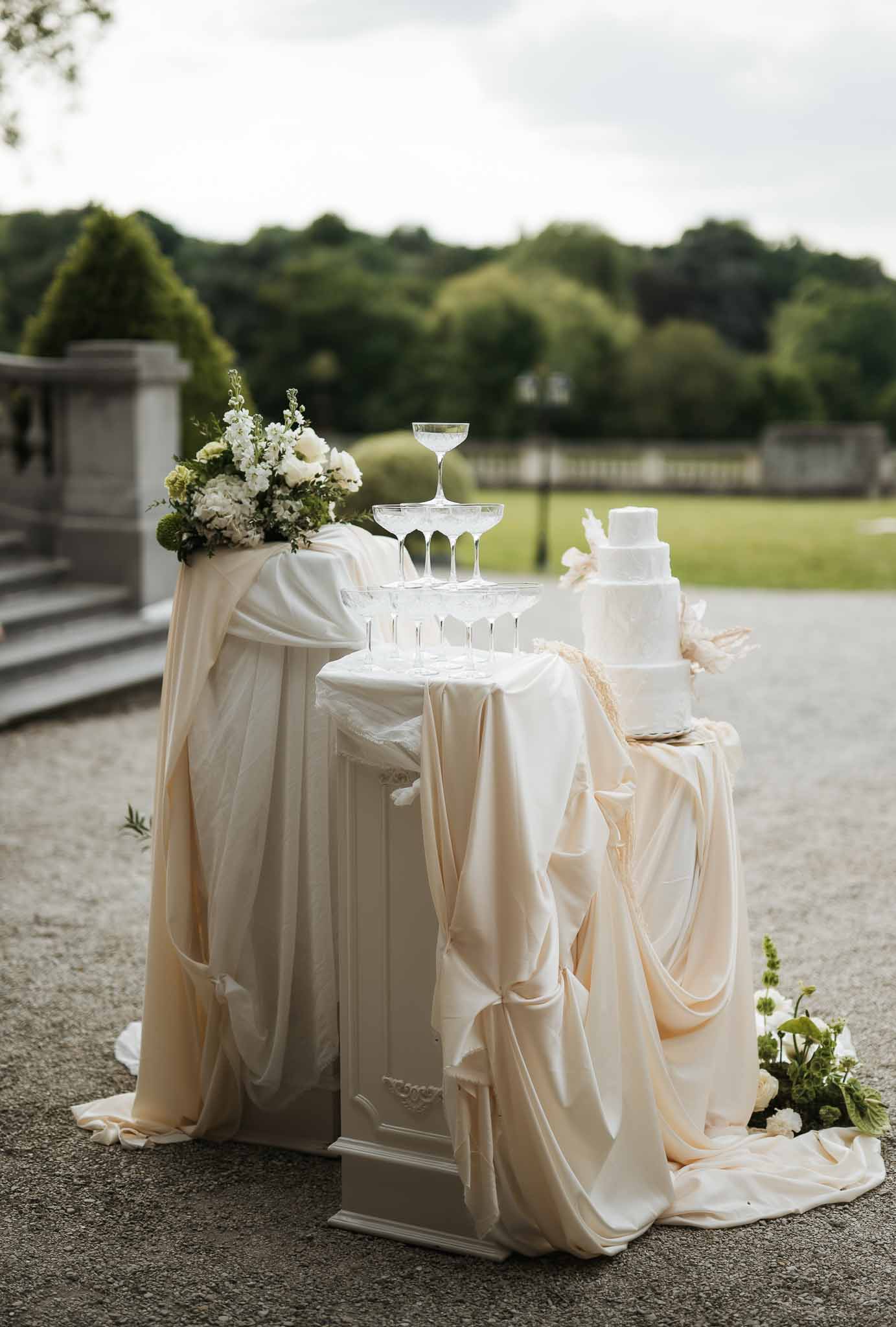 Three-tier white cake with pampas grass, hydrangea pedestal, and champagne coupe tower on blush fabric