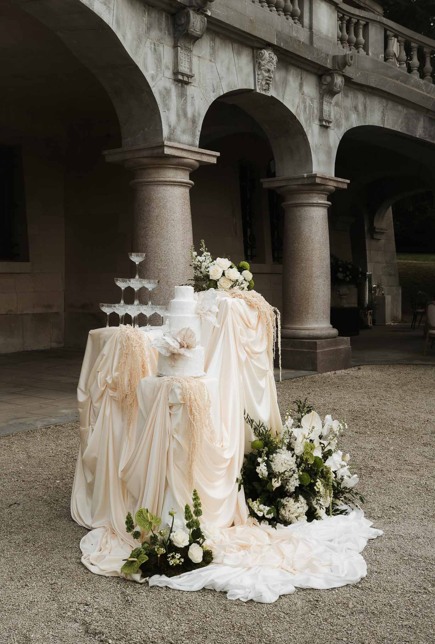 Multi-tier white cake with magnolia beside champagne tower and white rose installation before stone arcade