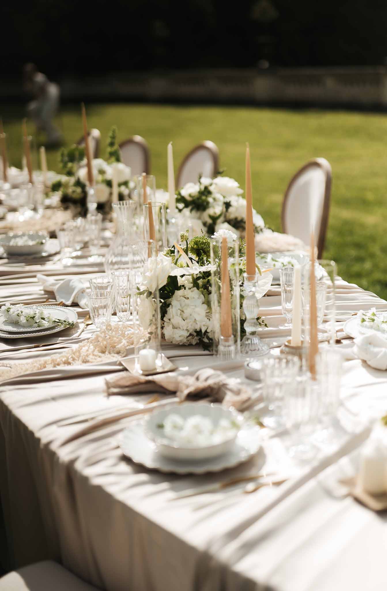 Outdoor reception table with white hydrangea centerpieces, gold flatware, crystal glassware, and tapered candles on greige...