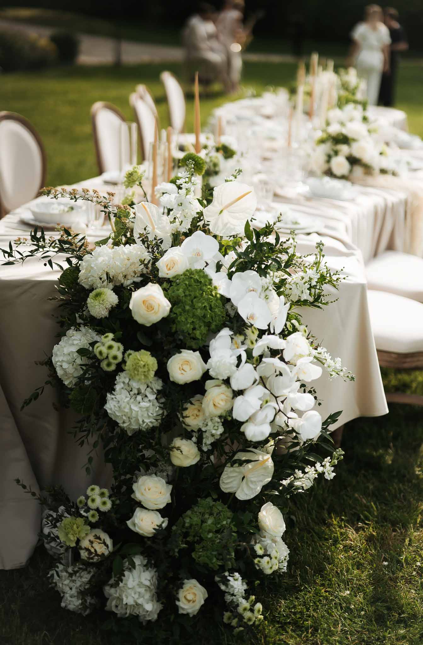 Cascading white orchid, rose, and anthurium centerpiece with gold taper candles on cream linen table