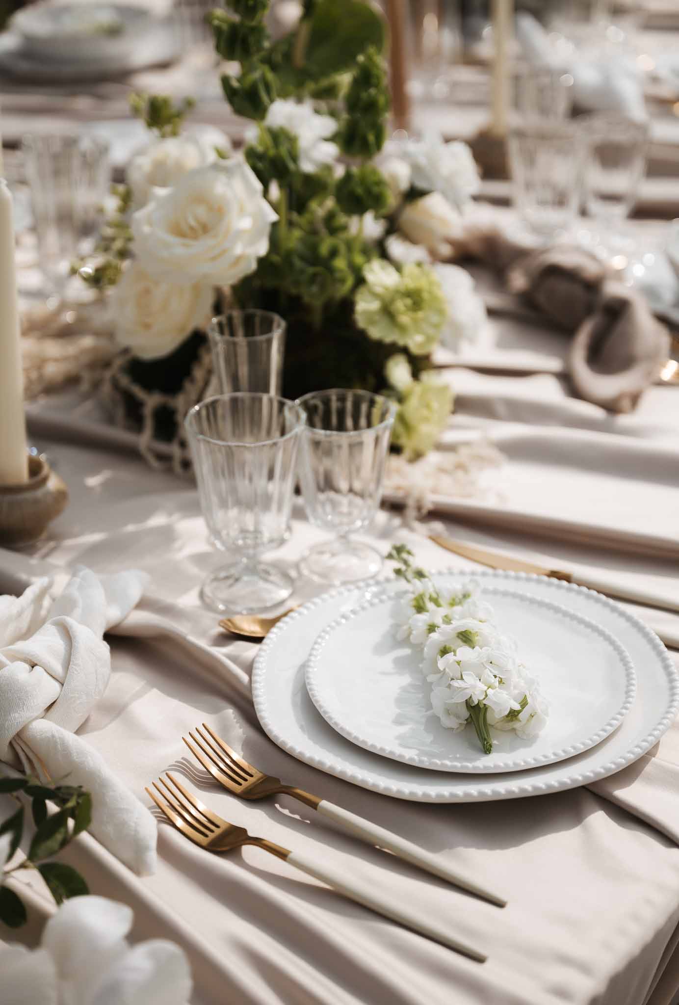 Reception place setting with white beaded plates, gold flatware, crystal glasses, and white floral centerpiece