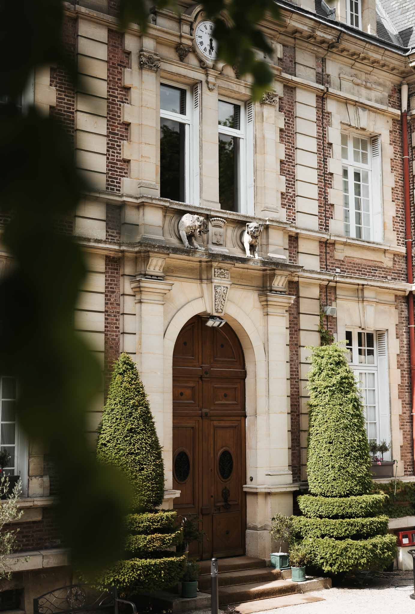 Red brick and limestone chateau facade with arched doors, topiary, and heraldic stone sculptures