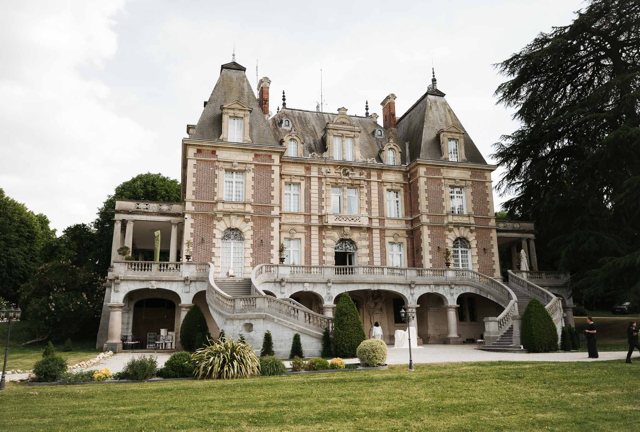 Grand red brick and limestone chateau with mansard roof, sweeping double staircase, and manicured topiary gardens
