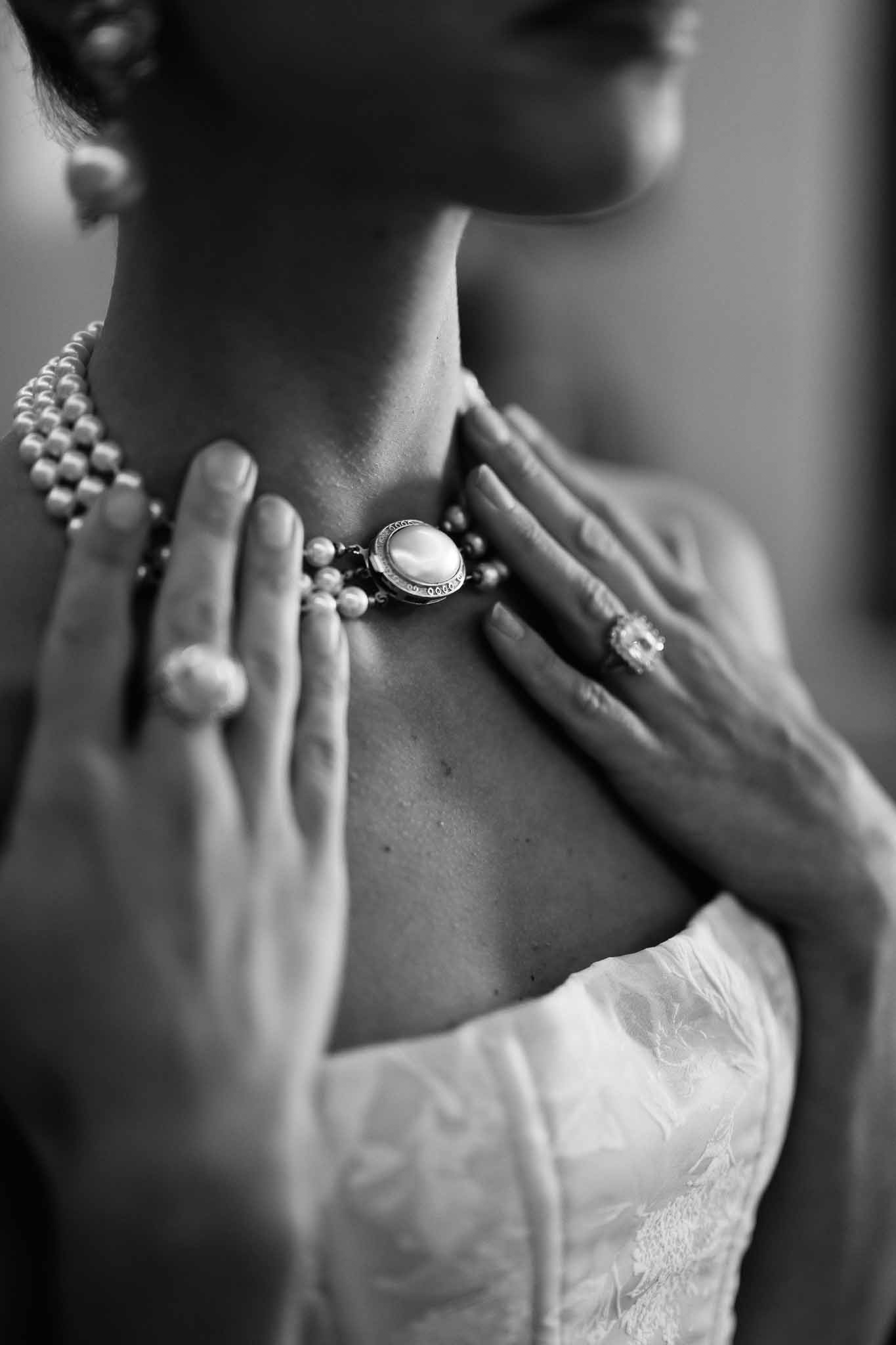 Black-and-white close-up of bride adjusting multi-strand pearl necklace with pearl and gemstone rings visible
