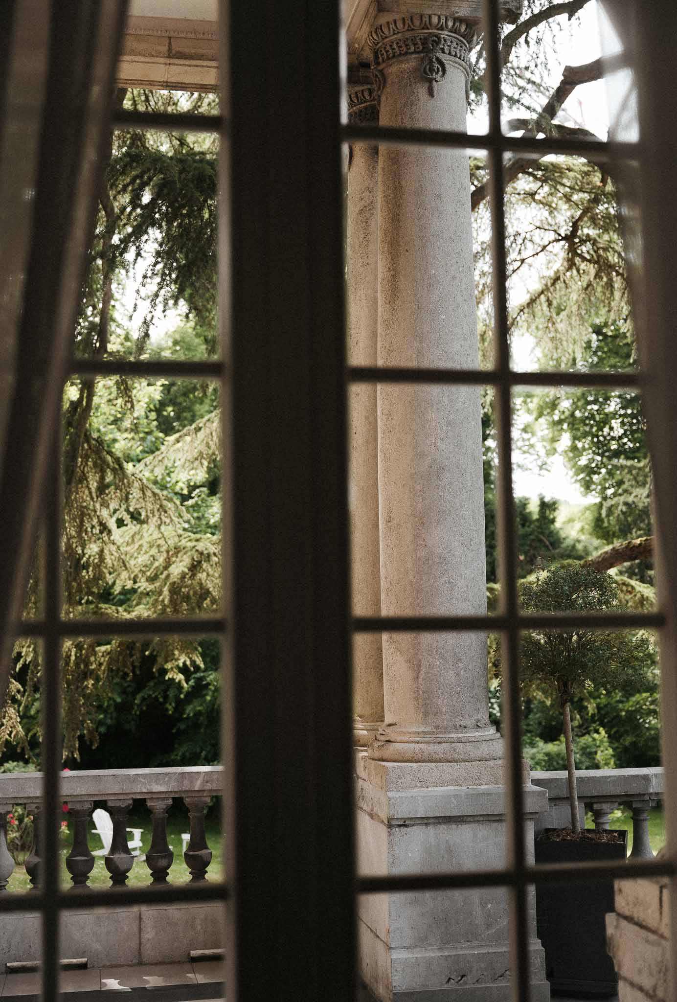 View through multi-pane window to terrace with classical stone column, balustrade, and potted topiary