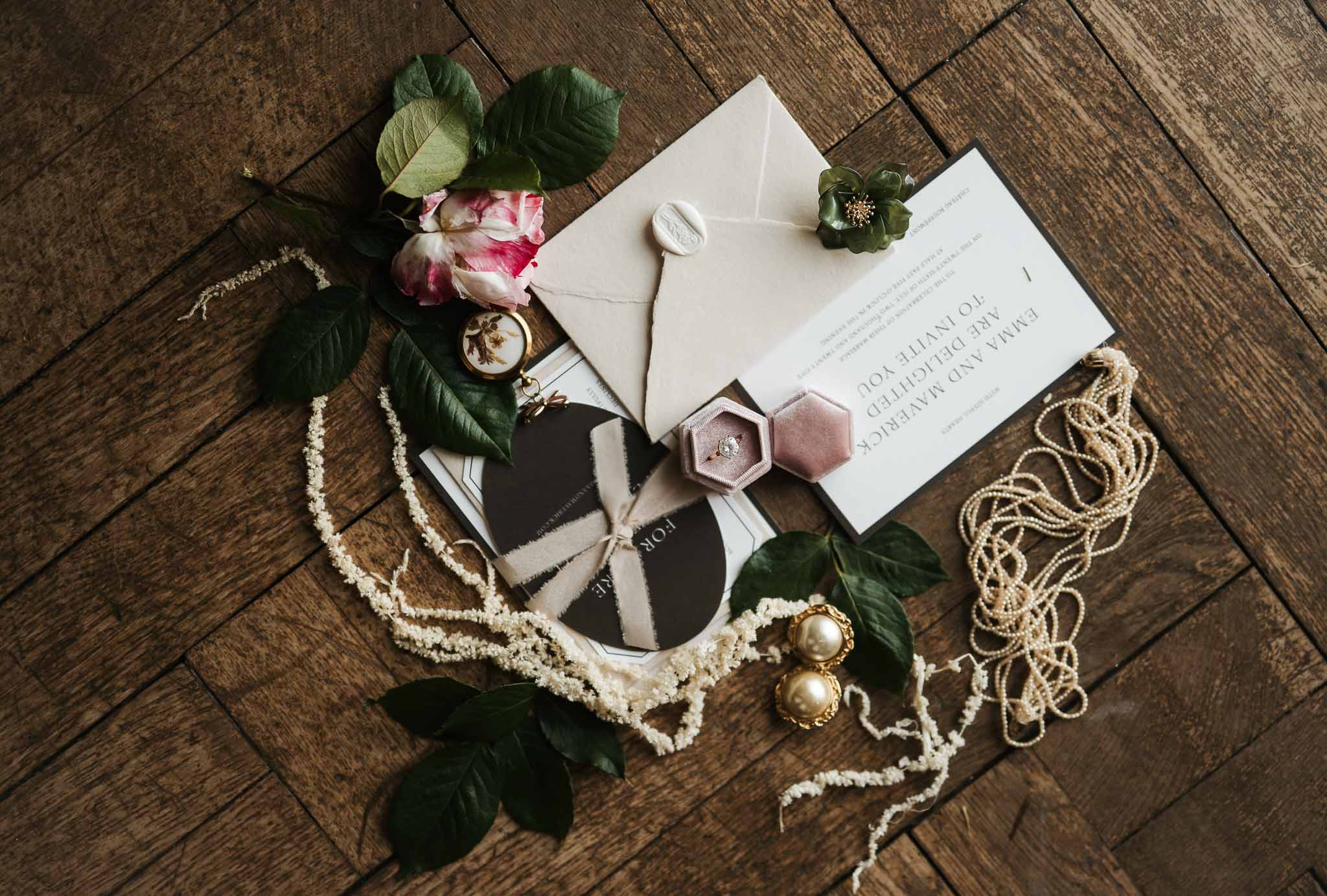 Flat lay of deckle-edge invitation with dusty mauve ring box, pearl strand, and pink garden rose