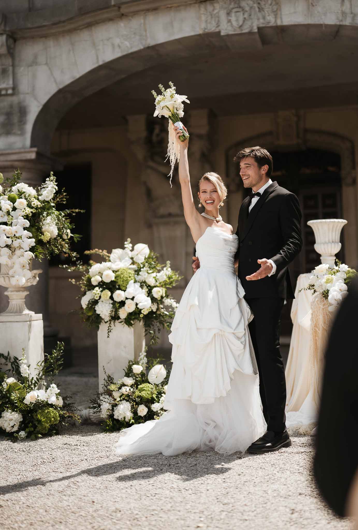 Bride and groom laughing in chateau courtyard after ceremony, bride holding white calla lily bouquet overhead