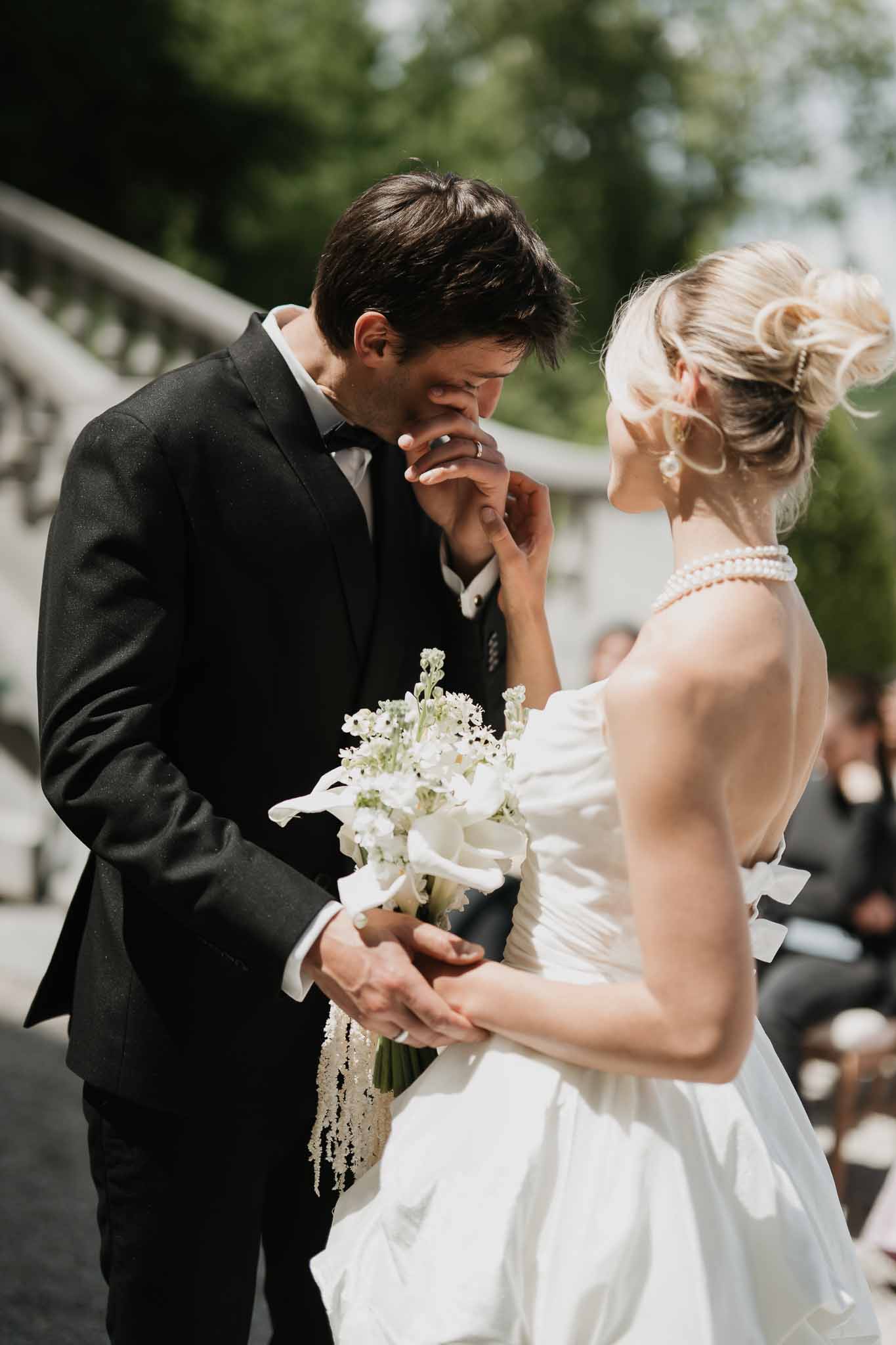 Groom kissing bride's hand as she holds white calla lily bouquet, pearl choker and stone balustrade behind